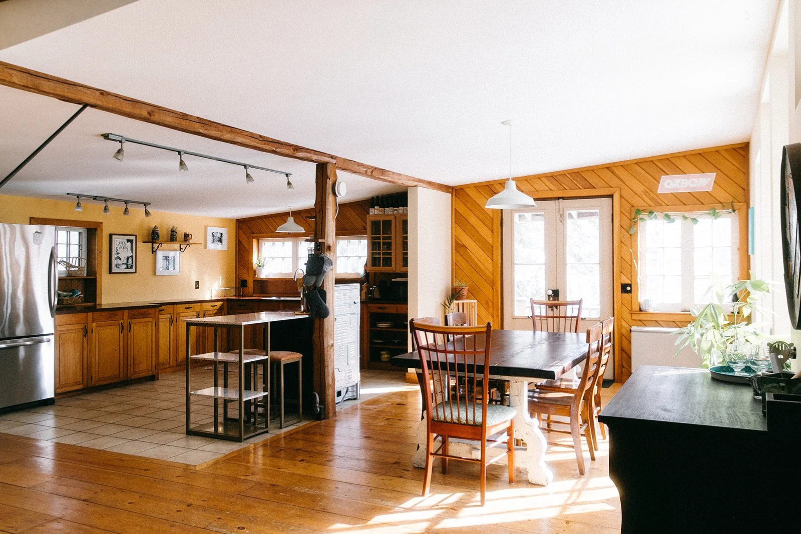 A cozy kitchen and dining area with wood accents, a tiled and wood floor, a stainless steel refrigerator, a hanging pot holder, a black table with wooden chairs, and large windows bringing in natural light.