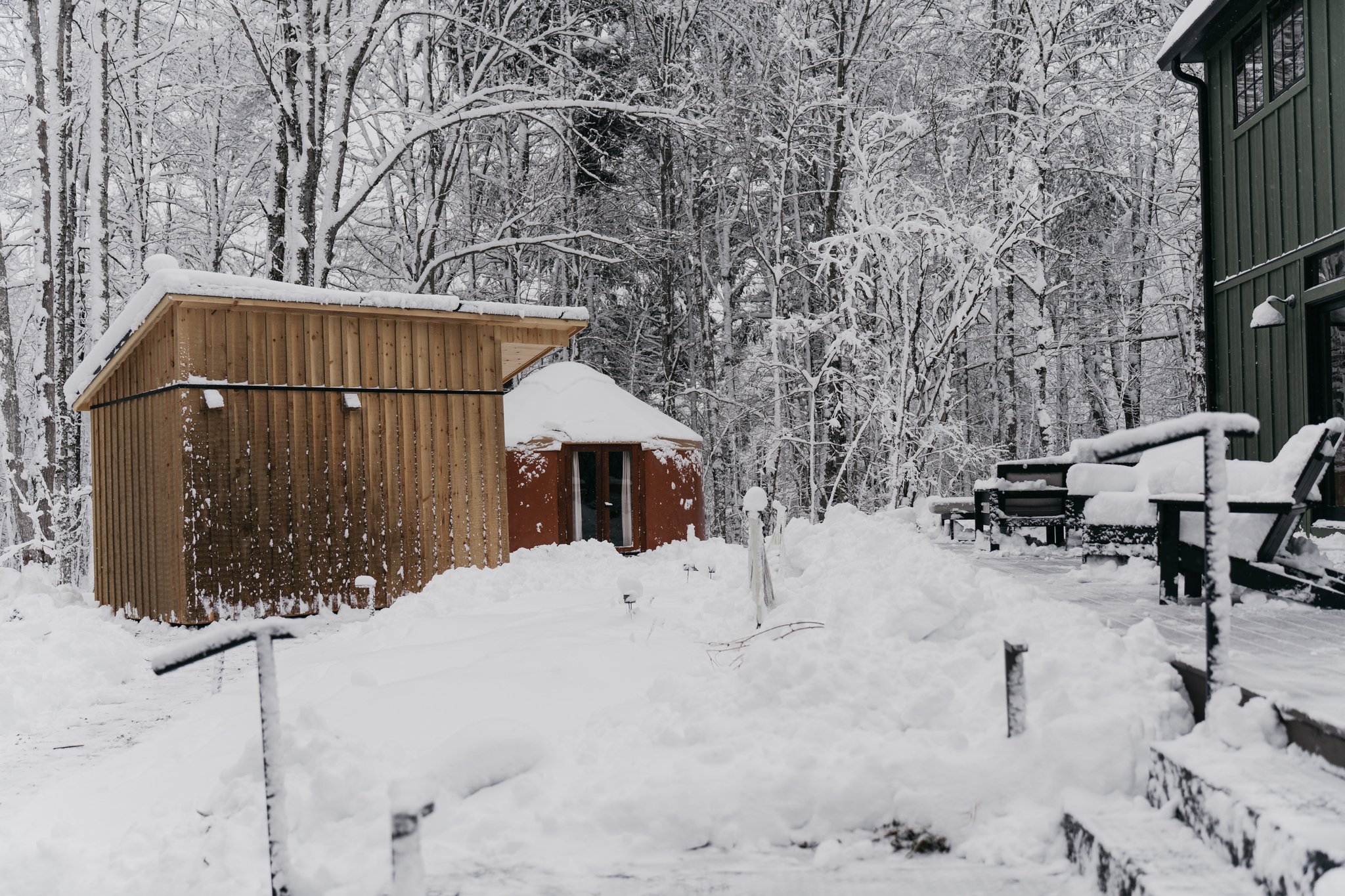 A snowy backyard with two wooden and one small green building, snow-covered trees in the background, and a snow-covered patio with outdoor chairs and benches.