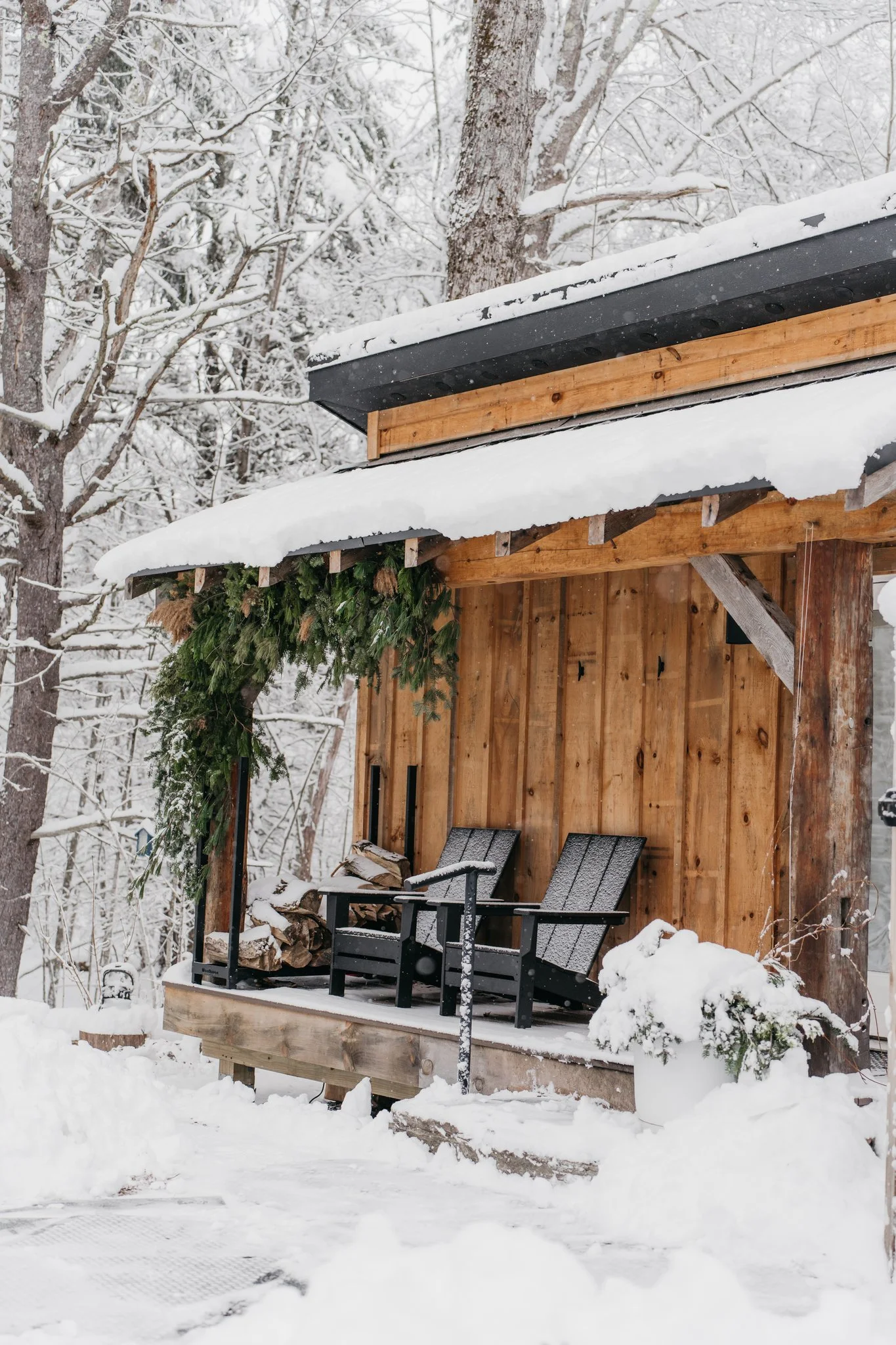 Snow-covered wooden porch with two black chairs, stacked firewood, greenery, and snow on the roof and ground.