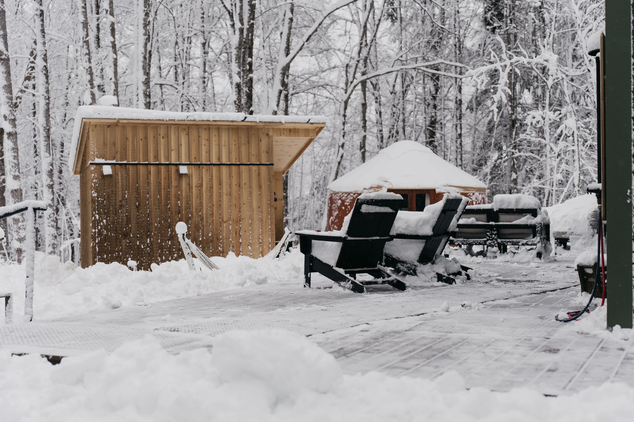 Snow-covered outdoor deck with wooden and black chairs, small shed, and trees covered in snow in the background.