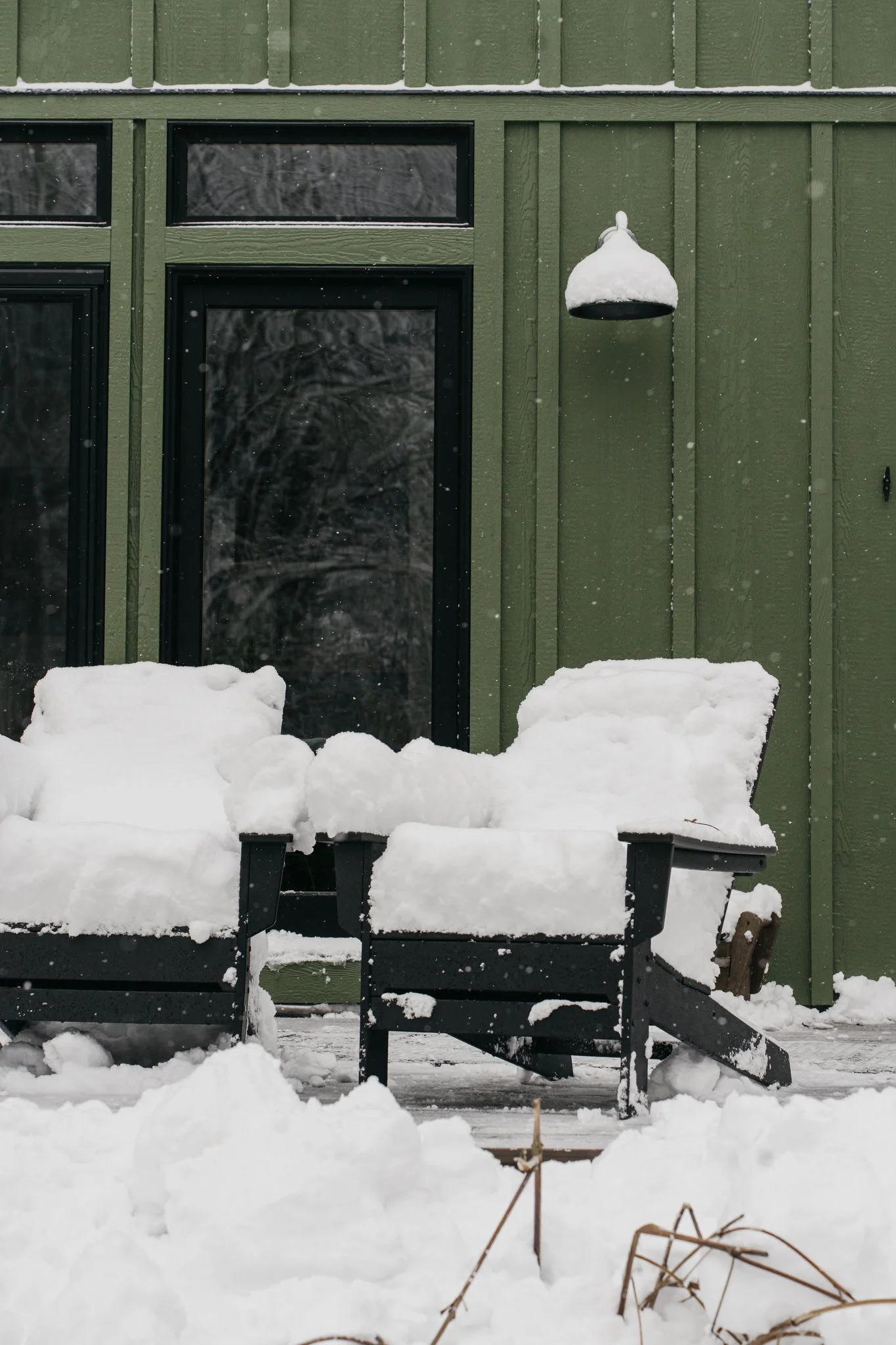 Snow-covered outdoor patio with two chairs, a side table, and a wall-mounted lamp in front of a green exterior wall, with snow falling.