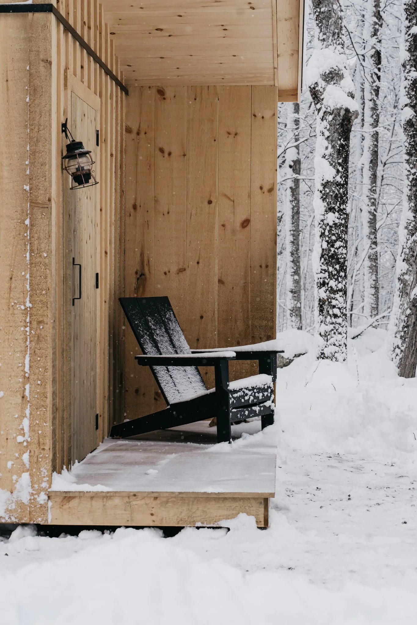 A black Adirondack chair covered in snow on a wooden porch, with a wooden wall and snow-covered trees in the background.