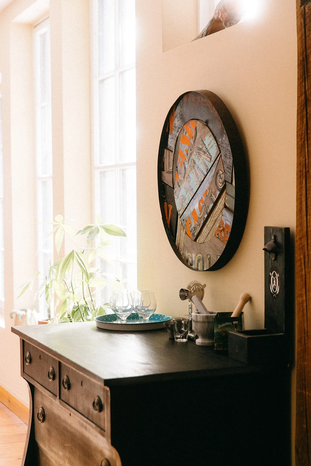 A wooden sideboard with glasses and a tray, a plant in front of a window, and a colorful round wall art.