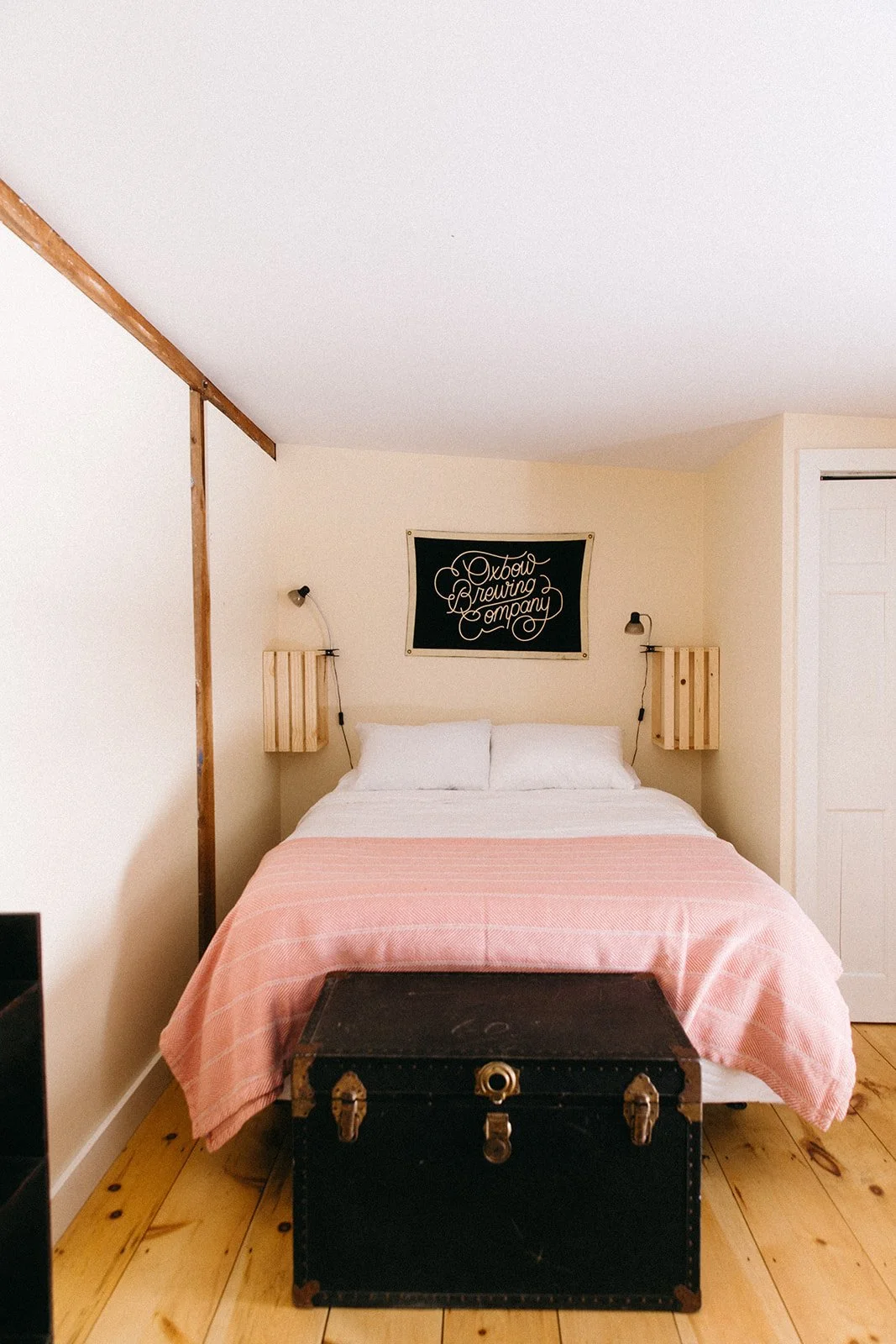 A bedroom with a bed, pink bedspread, white pillows, wooden nightstands with wall-mounted lamps, a sign that reads 'Oxbow Brewing Company', and a vintage black trunk at the foot of the bed.