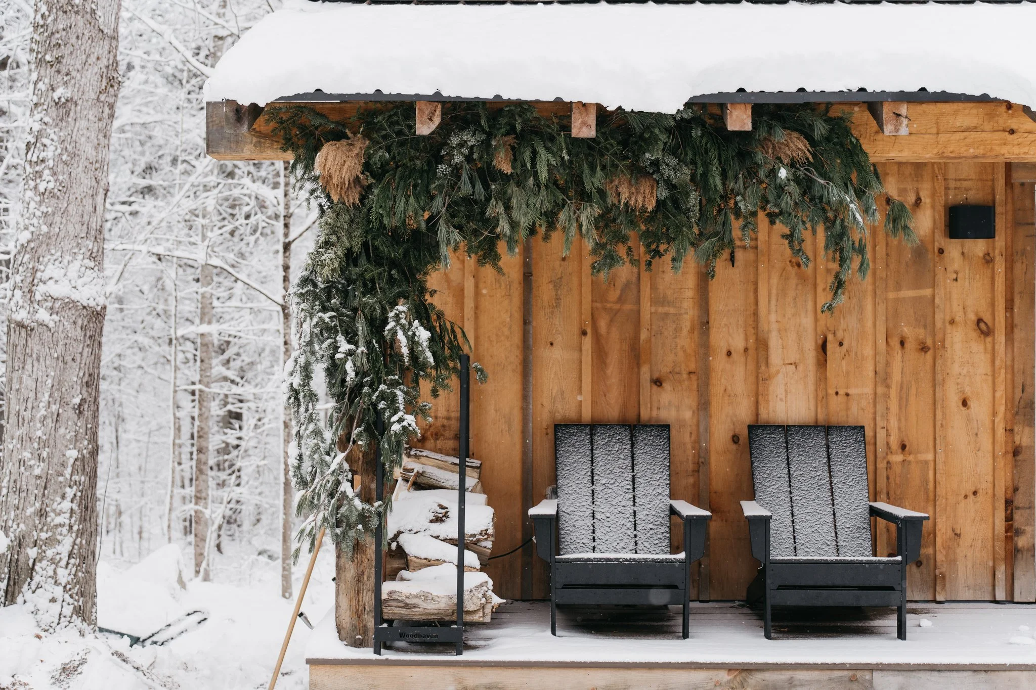 A wooden outdoor deck with two black chairs covered in snow, a pile of firewood, and a snow-covered canopy decorated with green garland, set in a snowy forest.
