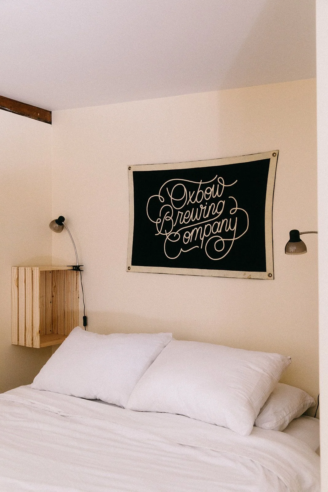 A bedroom with a bed, white bedding, and pillows. A wall-mounted wooden shelf and two wall lamps on either side. A black sign with white cursive text reading 'Oxbow Brewing Company' hangs above the bed.
