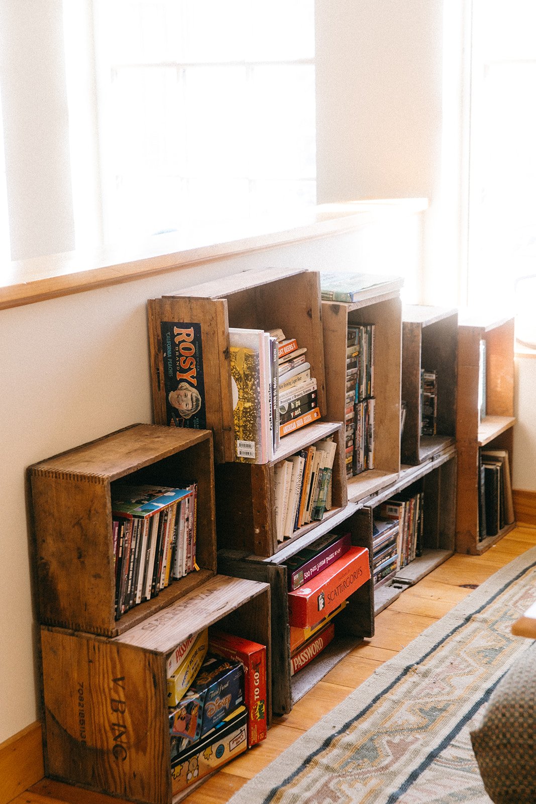 Wooden bookshelves filled with books and board games against a wall, with a window and a colorful rug on wooden flooring.