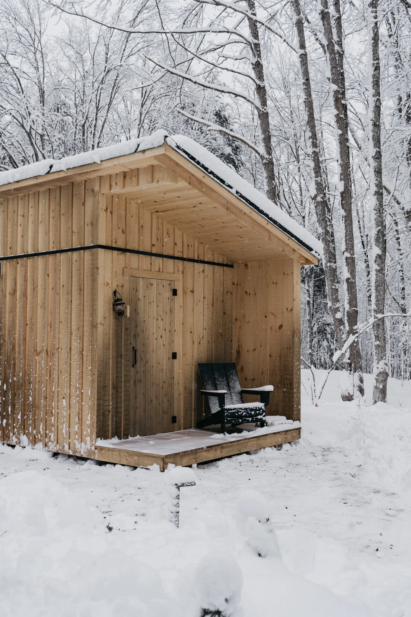 A small wooden cabin on a snowy ground, with snow on the roof and a black rocking chair on the porch, surrounded by snow-covered trees.