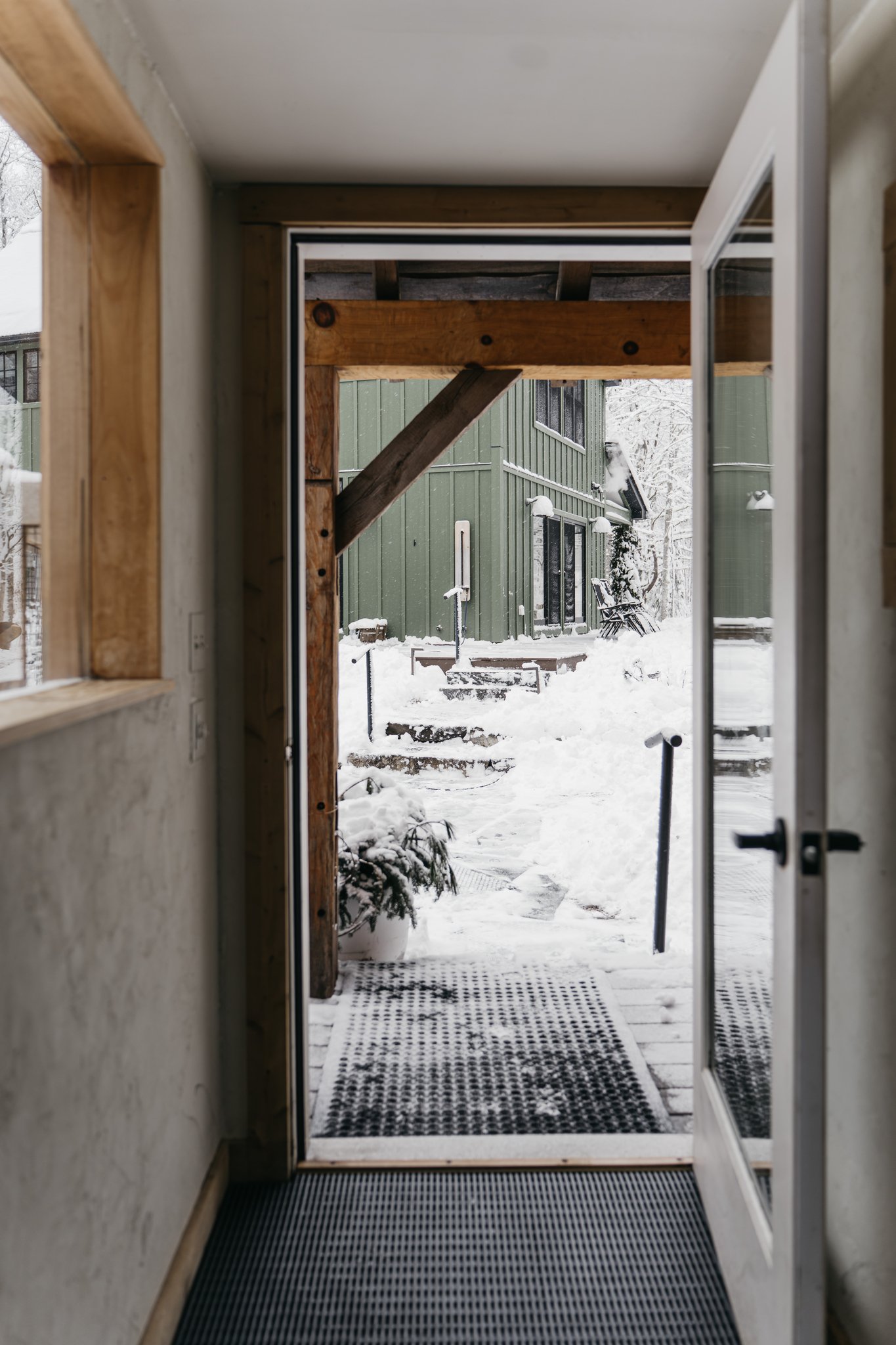 View through an open door showing a snowy outdoor scene with a green building, steps covered in snow, and outdoor furniture.