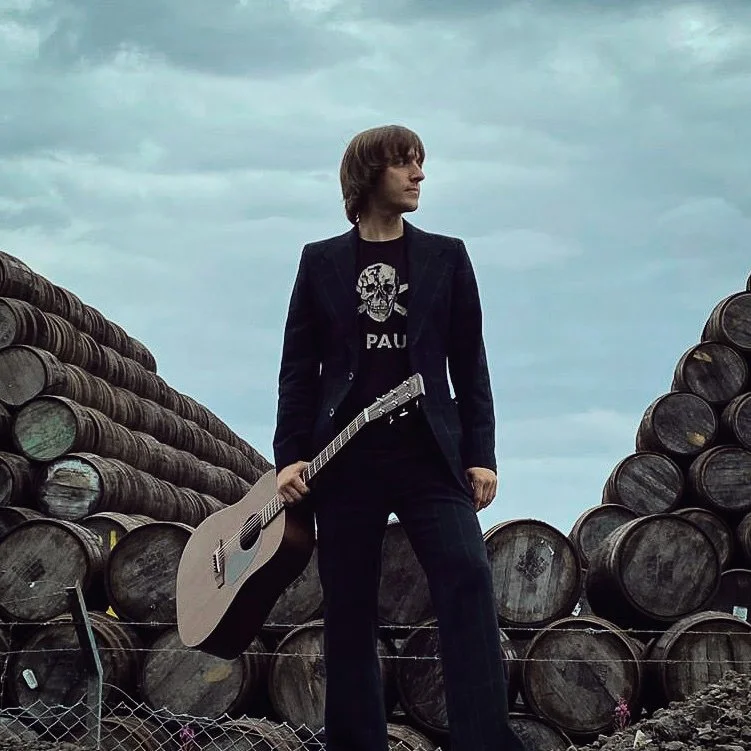 A woman standing outdoors between stacks of large wooden barrels, holding an acoustic guitar, under cloudy sky.