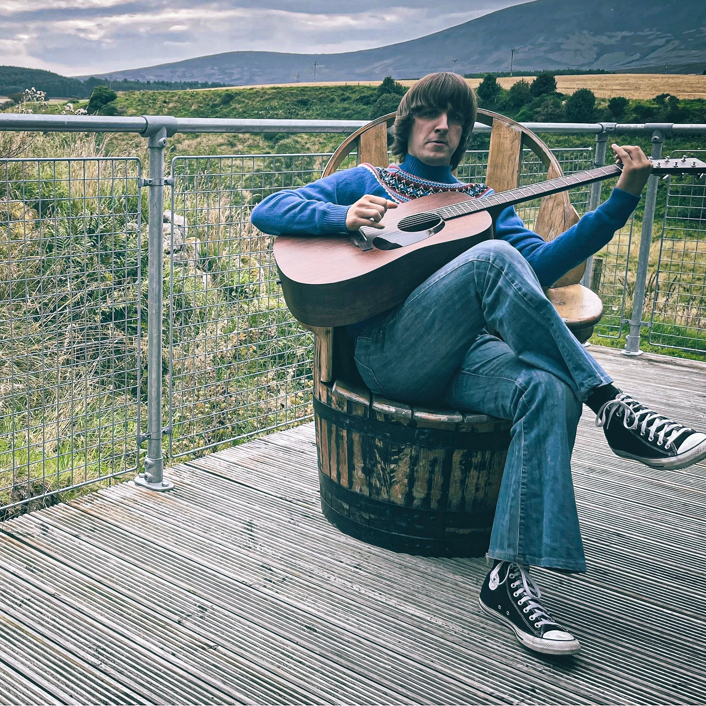 A person sitting on a wooden barrel on a metal deck, playing an acoustic guitar, with a mountain landscape in the background.
