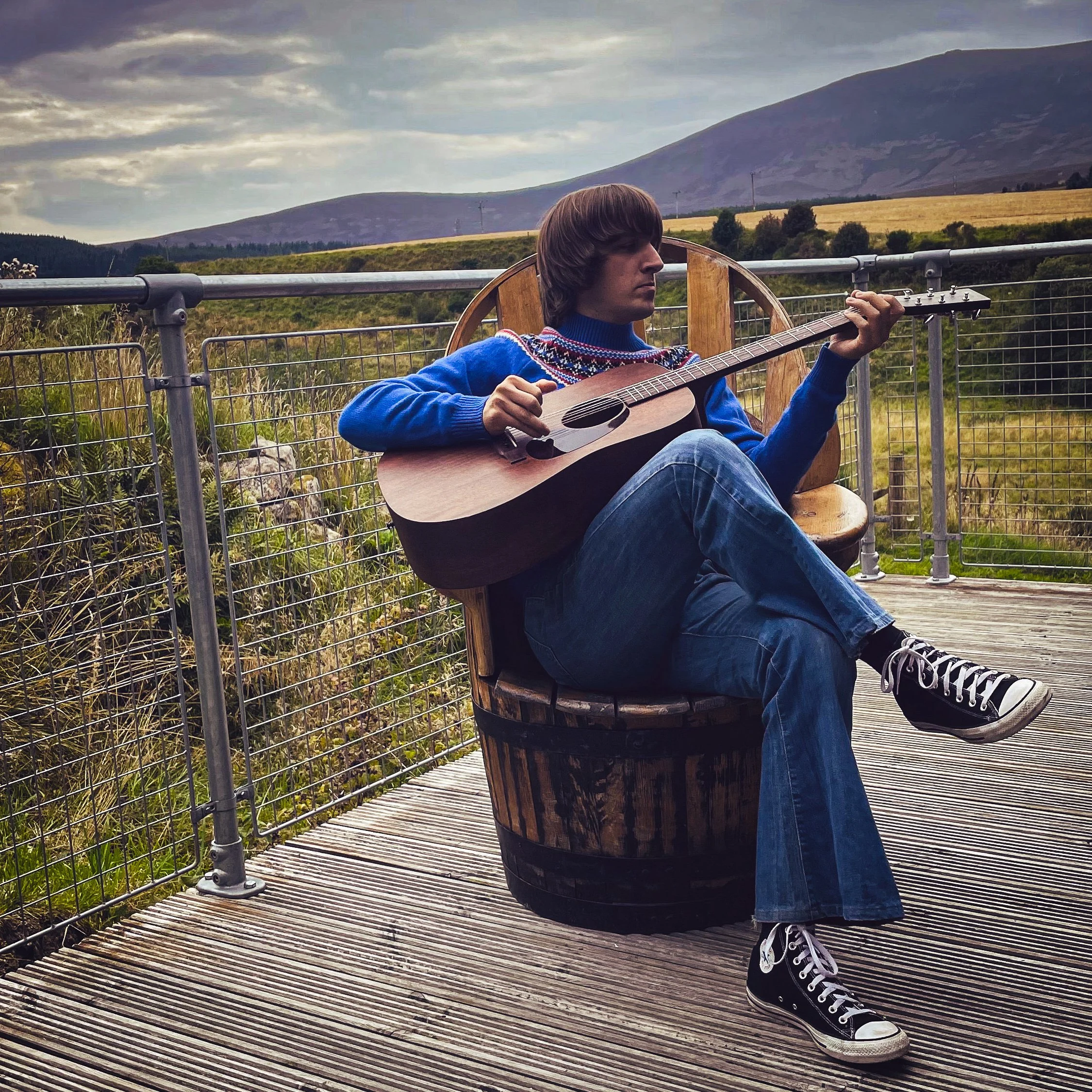 Person sitting on a wooden bench on a bridge, playing an acoustic guitar with a mountainous landscape in the background.