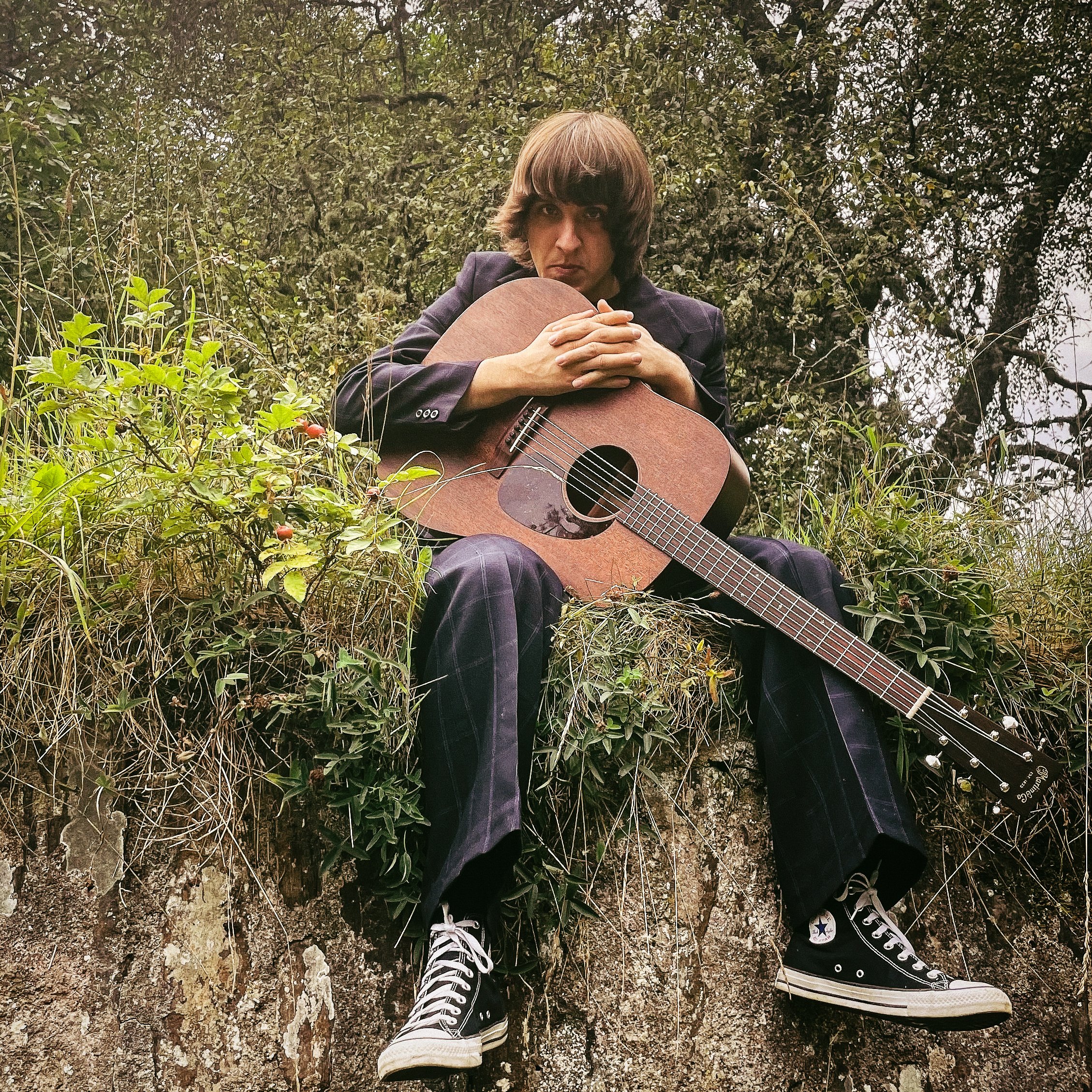 Person sitting outdoors on a rocky surface, holding a guitar, surrounded by greenery and shrubs, wearing plaid pants and black Converse shoes.