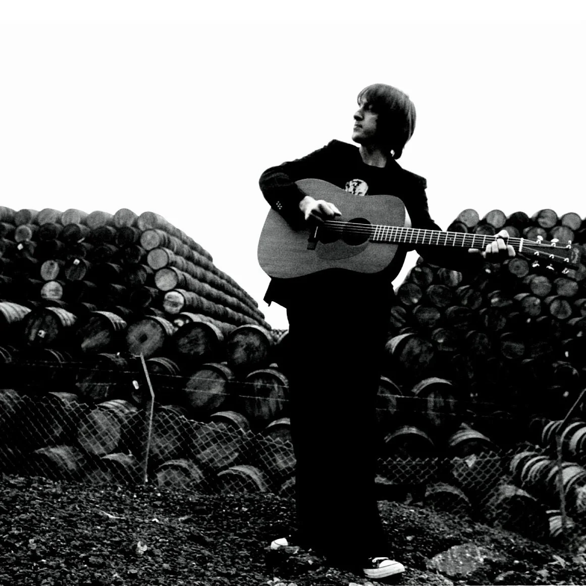 A person playing an acoustic guitar outdoors, standing in front of stacked logs, in black and white.