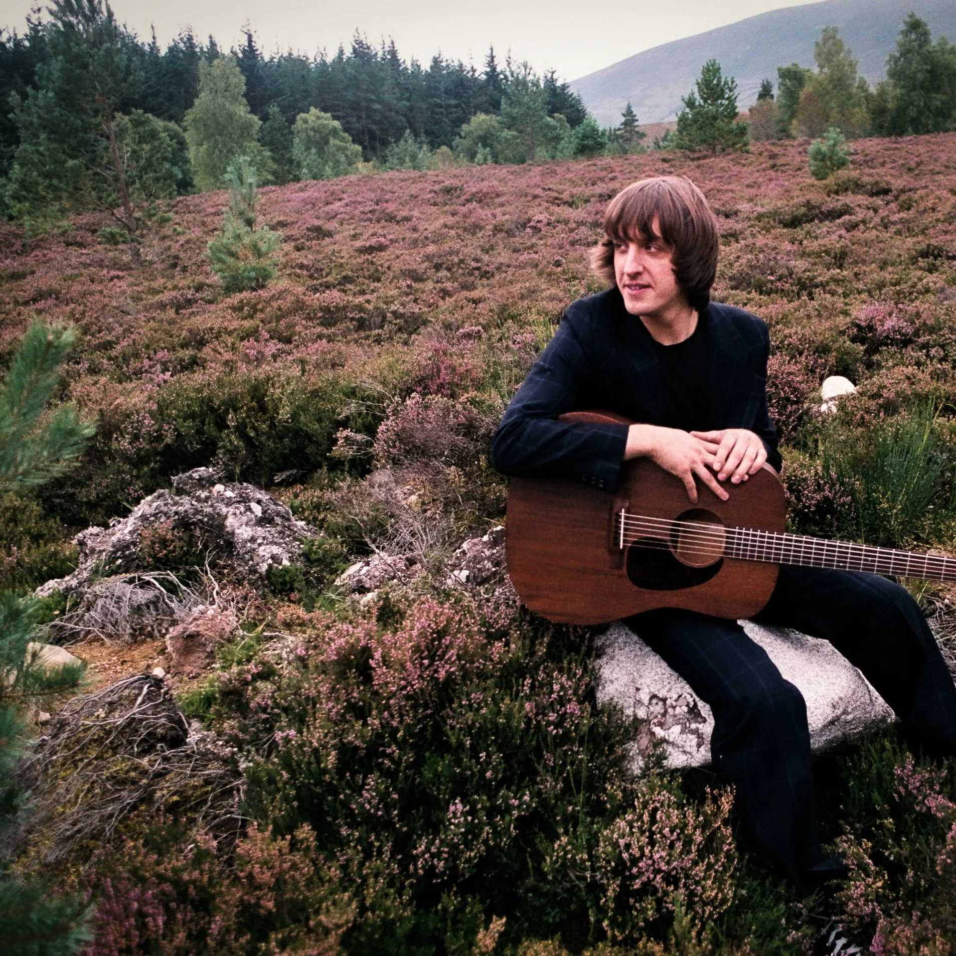 A young man with brown hair sitting in a field of pink heather with green trees and mountains in the background, holding a guitar.
