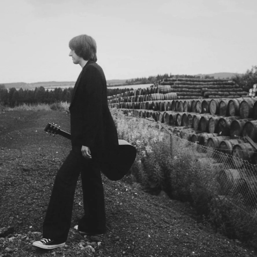A woman standing outdoors on a dirt path, holding an acoustic guitar, with rolls of hay in the background.