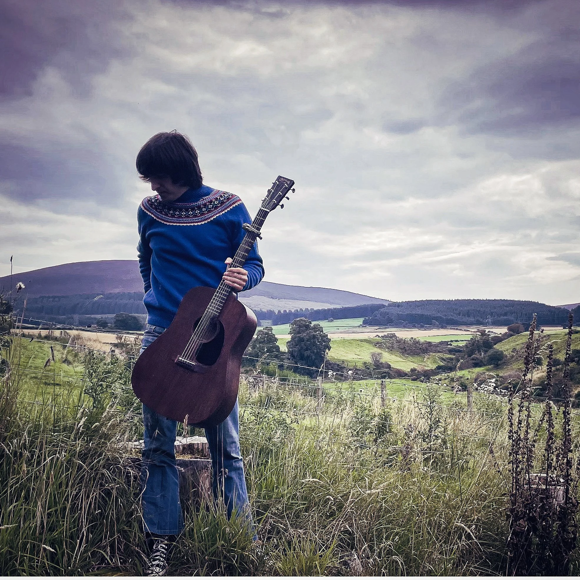 A person with shoulder-length dark hair wearing a blue sweater with a decorative pattern on the shoulders is holding an acoustic guitar in a landscape with grassy fields and distant hills under a cloudy sky.