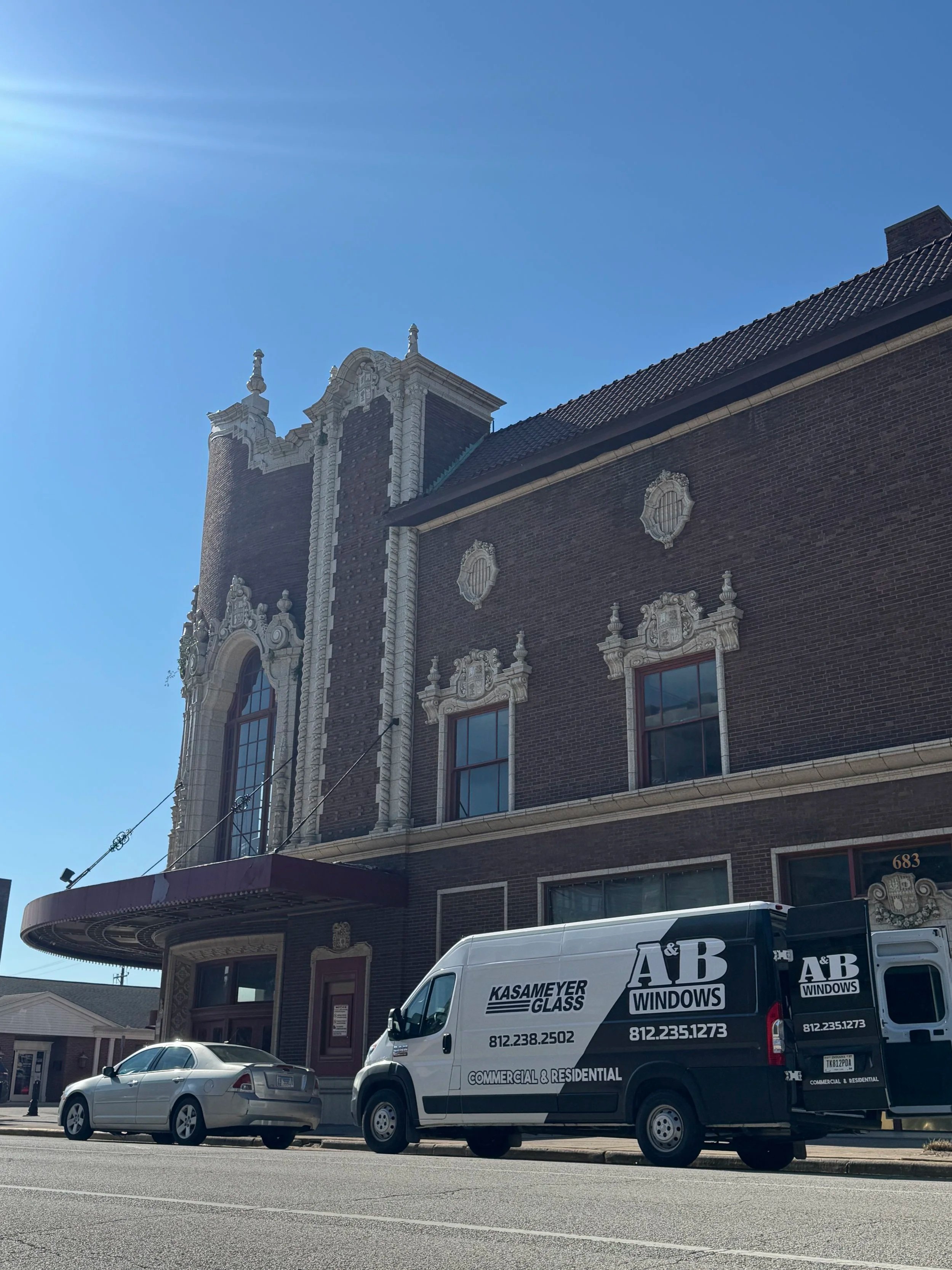 A street view featuring a historic brick building with ornate white decorative trim and a tower, parked cars, and a white commercial van with black lettering advertising glass and window services. The sky is clear and blue.