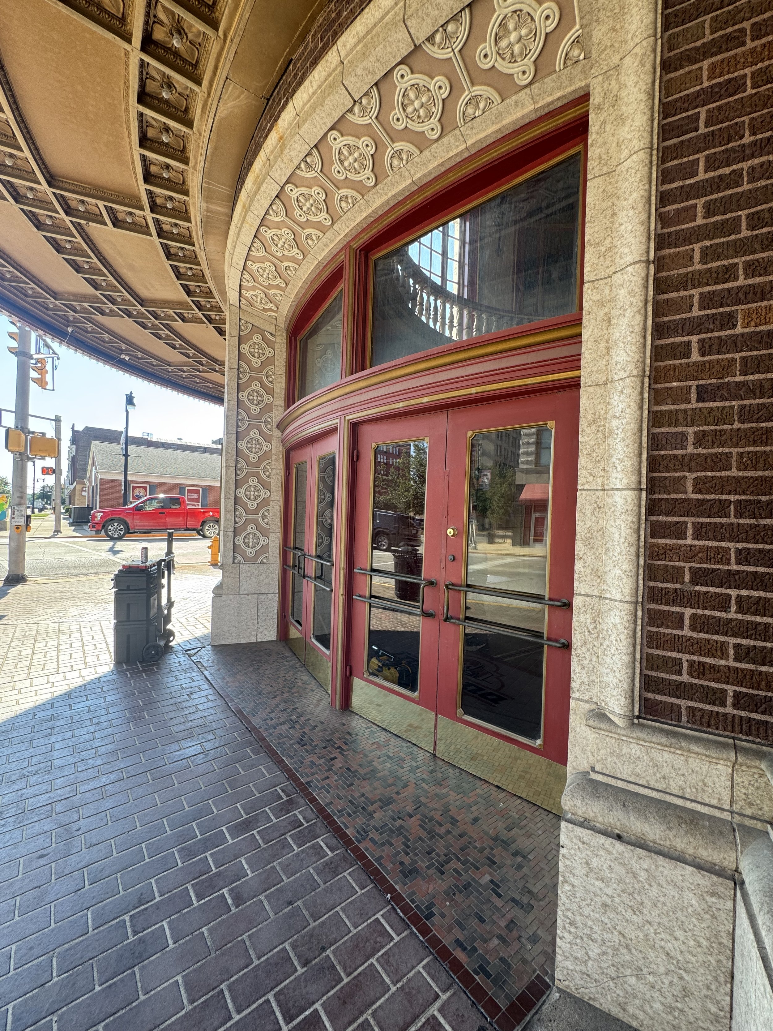 Exterior of a building with a decorative arch, pink double doors with glass panels, intricate patterns, and brick wall. There is a sidewalk, a street with a red truck, and streetlights visible in the background.