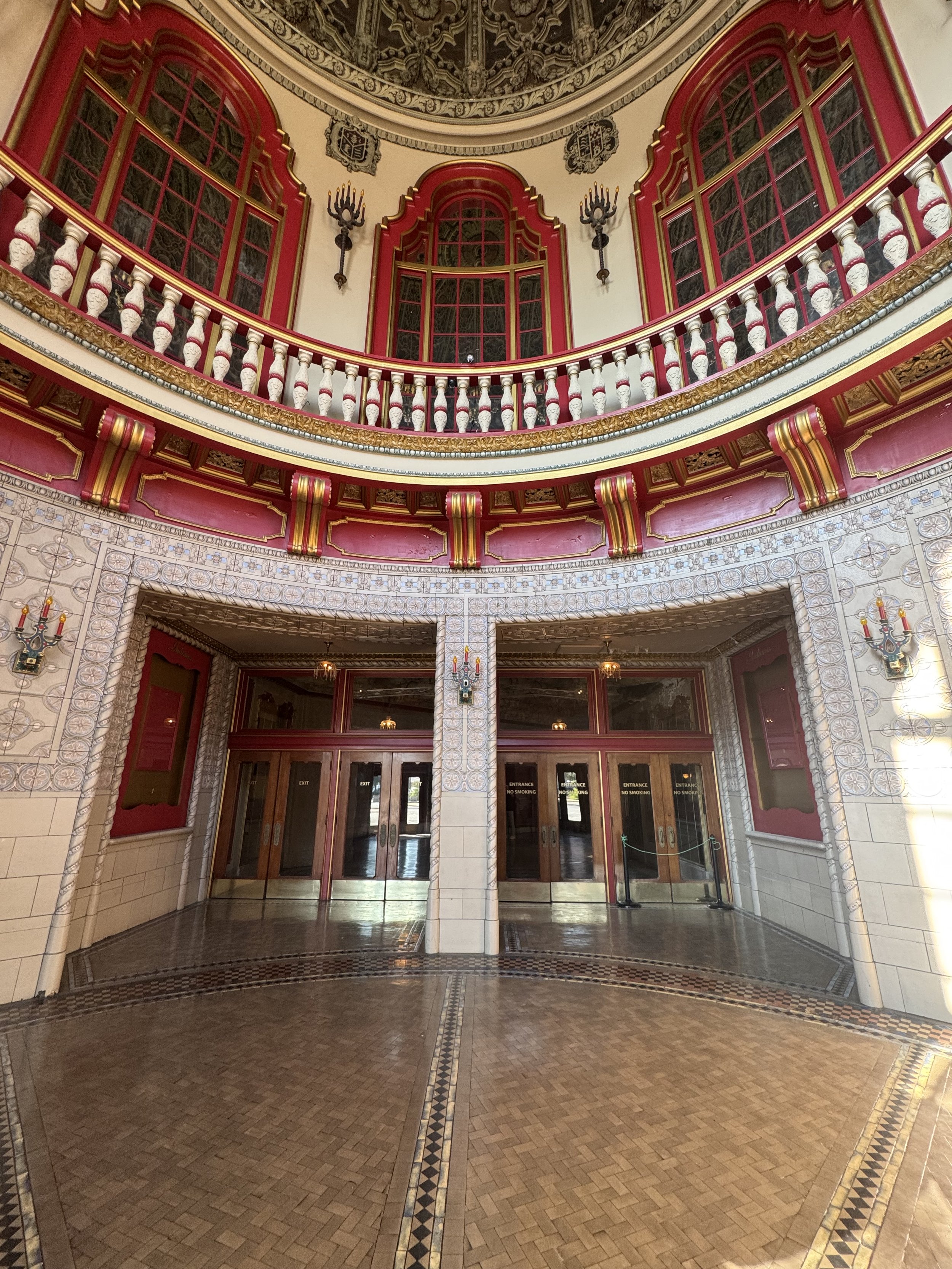 Elegant interior with a grand archway, ornate red and gold accents, and decorative wall sconces, leading to a hallway with wooden floors.