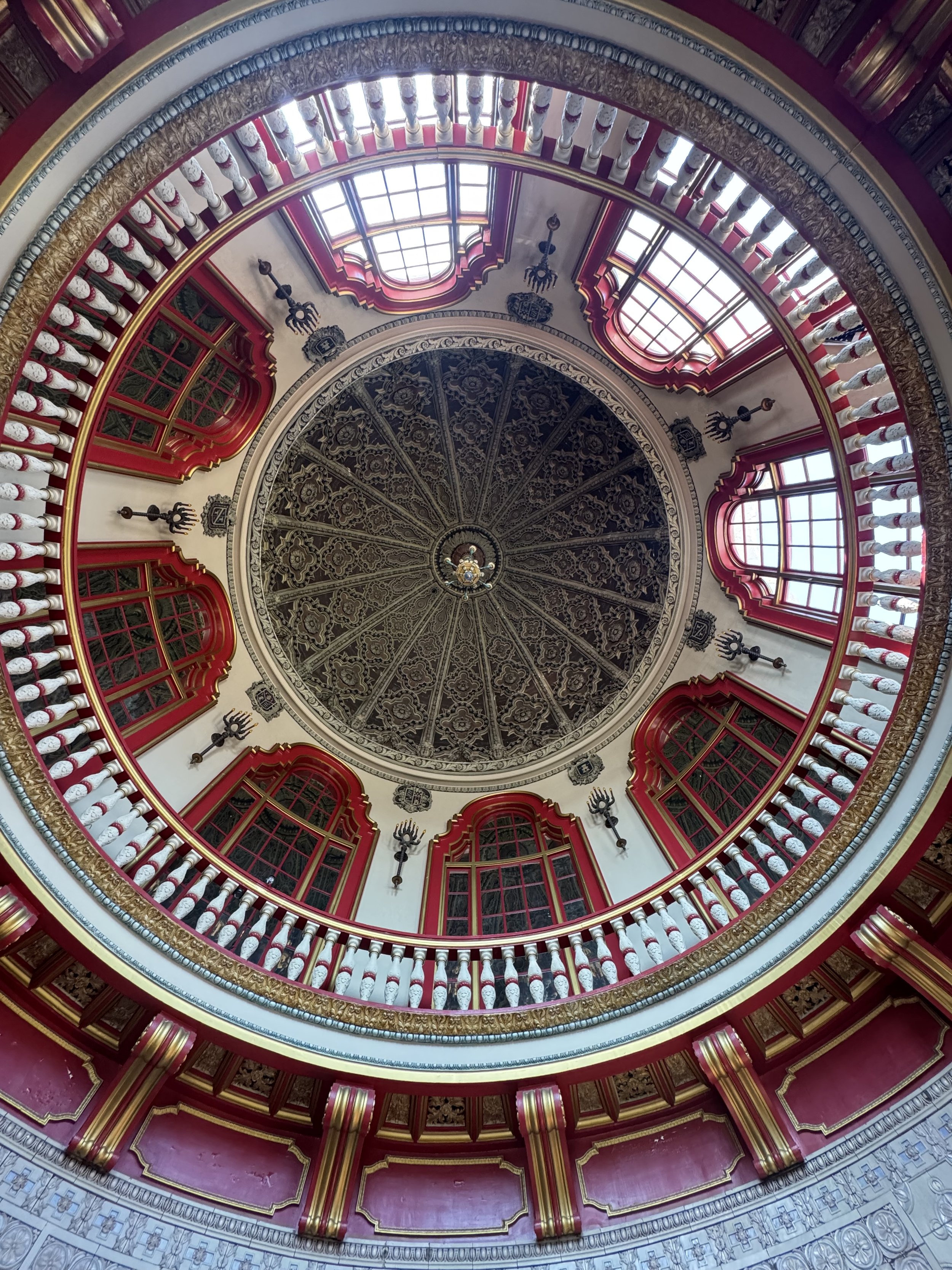 Interior view looking up at a domed ceiling with ornate detailing, surrounded by windows and red and gold decorative architectural elements.