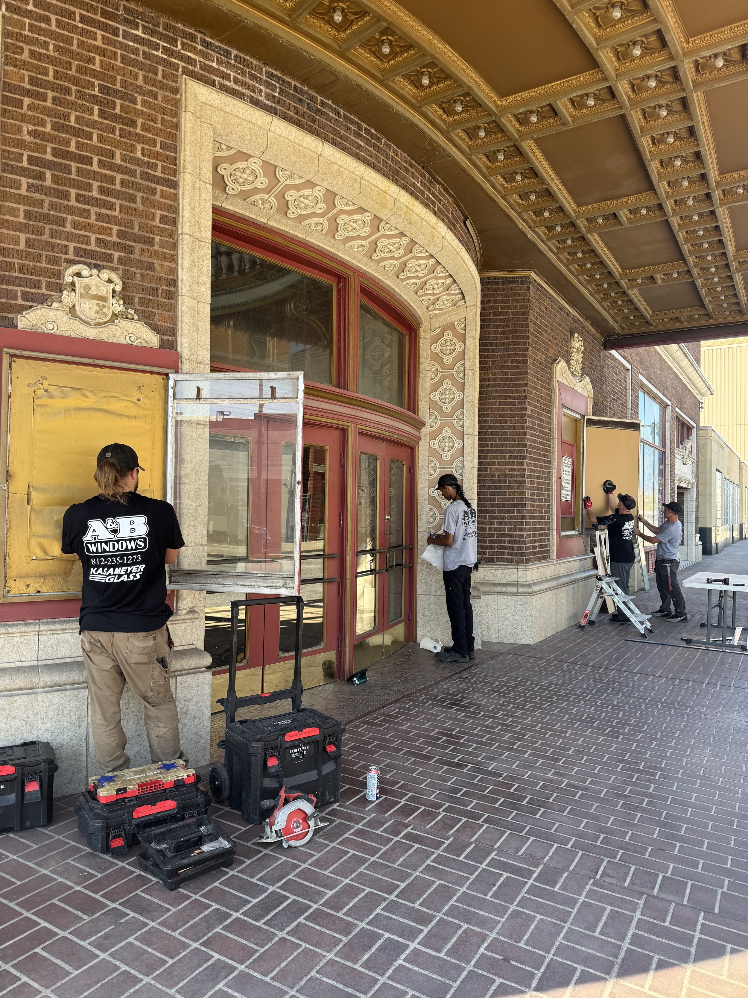 Workers setting up a building's exterior, with tools and equipment on the ground including power drills and a circular saw, outside a red brick building with decorative architectural details and large windows.