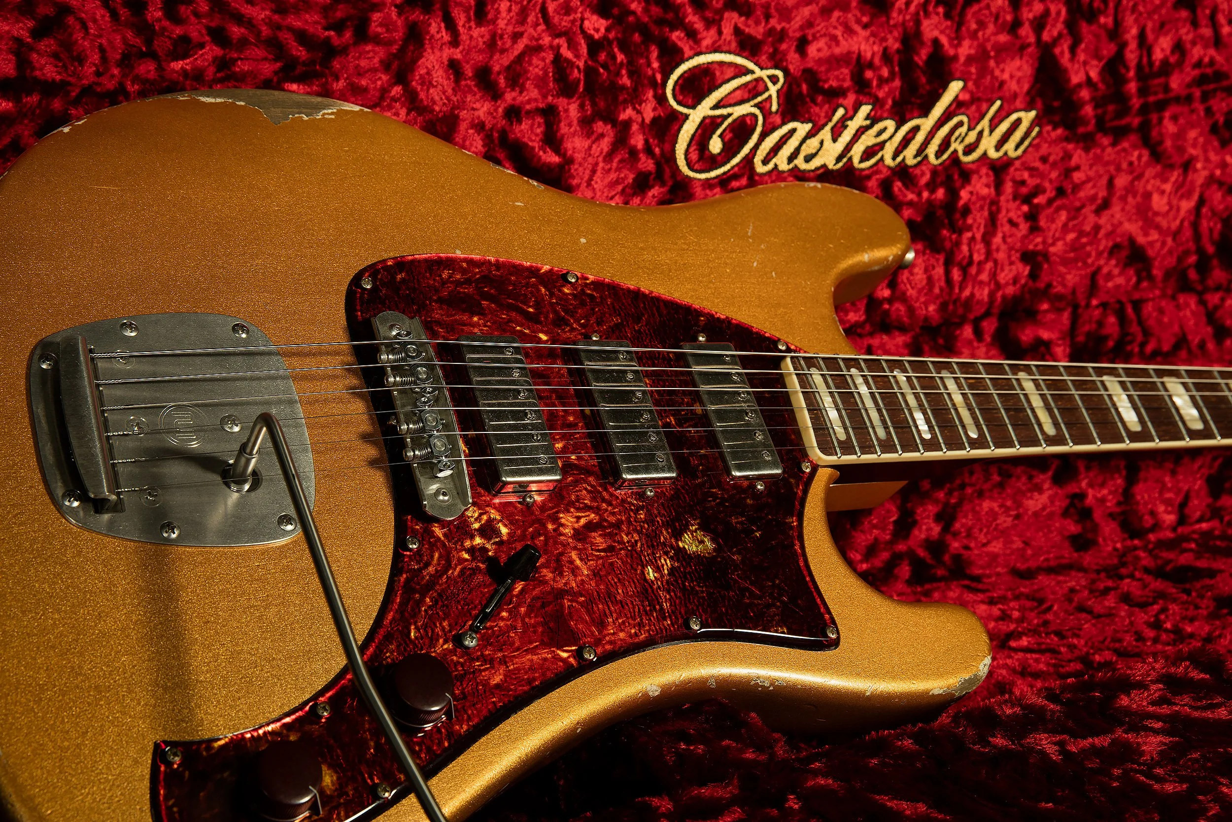 A gold electric guitar with a dark red tortoiseshell pickguard, set against a red velvet background with the word 'Castadosa' embroidered in white.