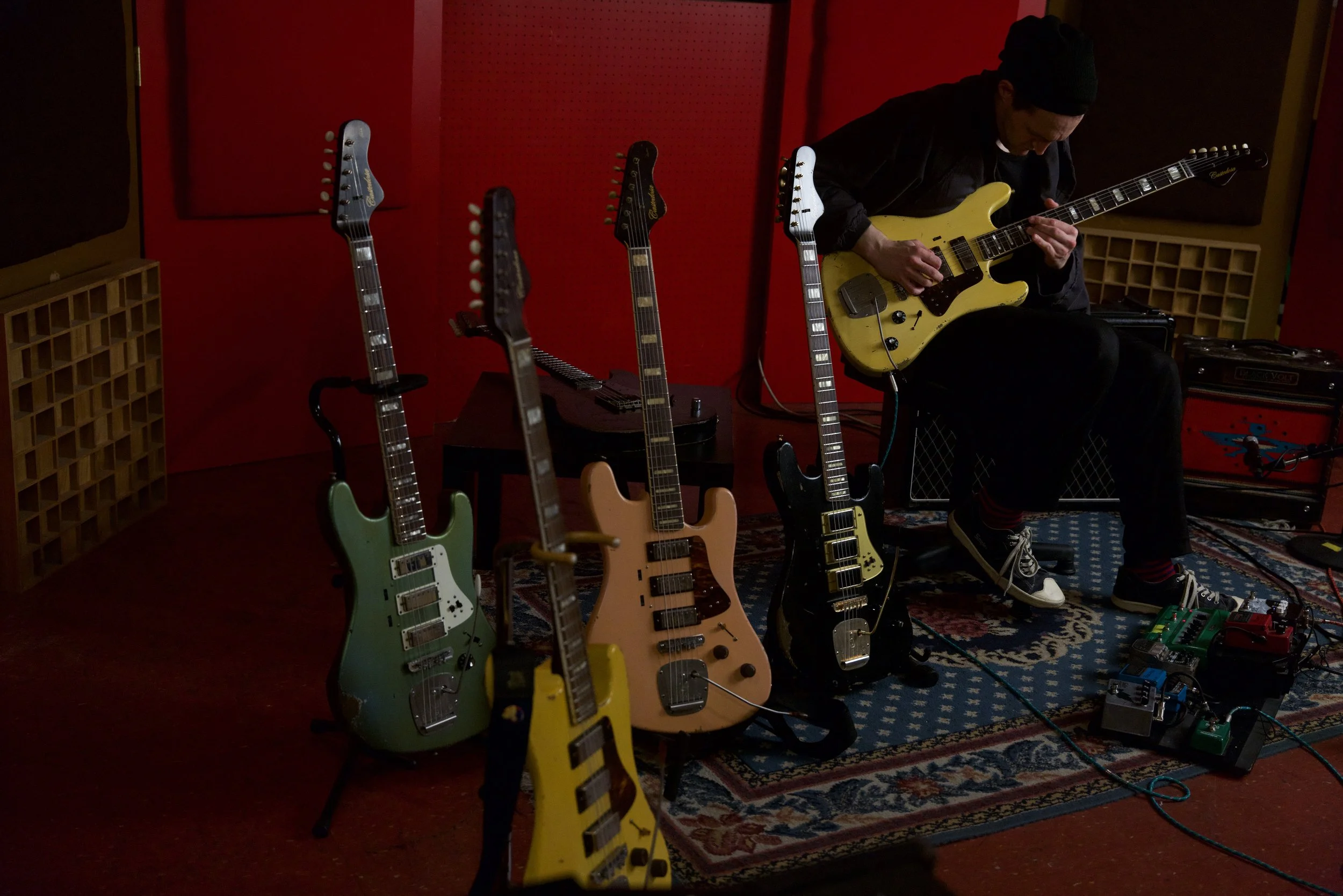 A man sitting on a stool playing a yellow electric guitar in a recording studio, surrounded by several other electric guitars on stands, with guitar effect pedals on the floor.