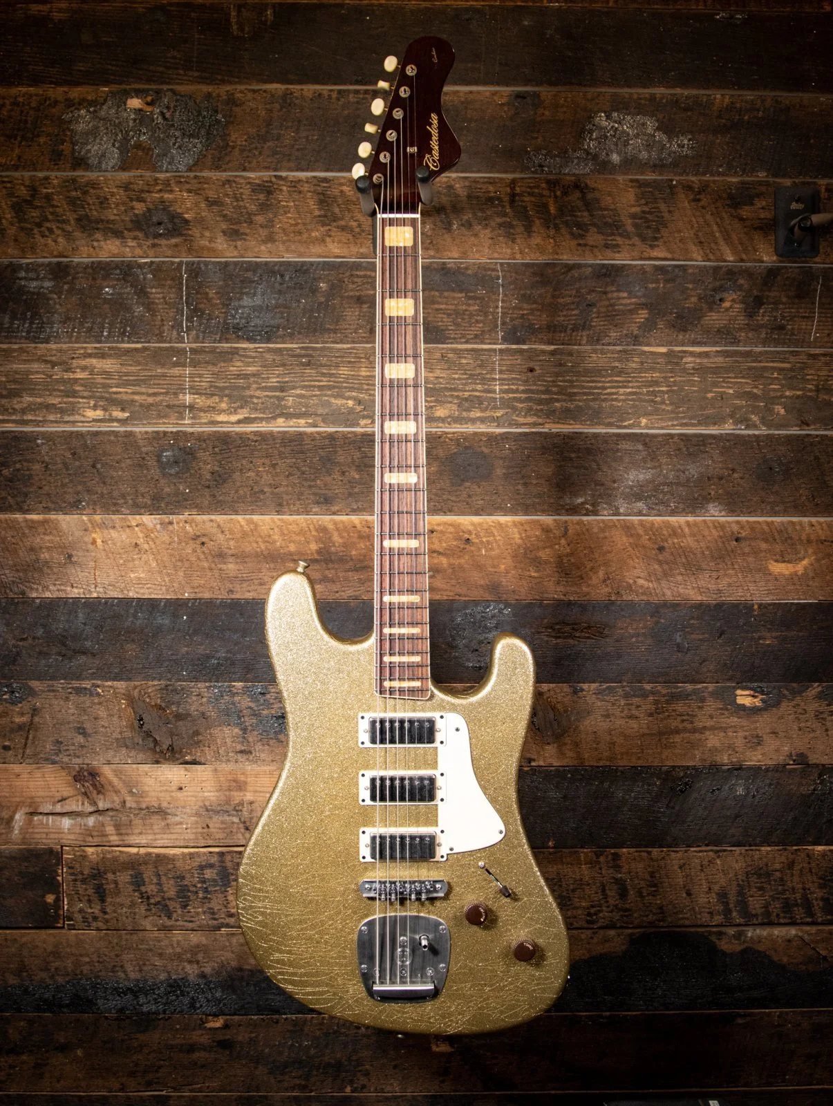 Gold electric guitar with a white pickguard, three pickups, and a crackled finish, on a dark wooden wall background.