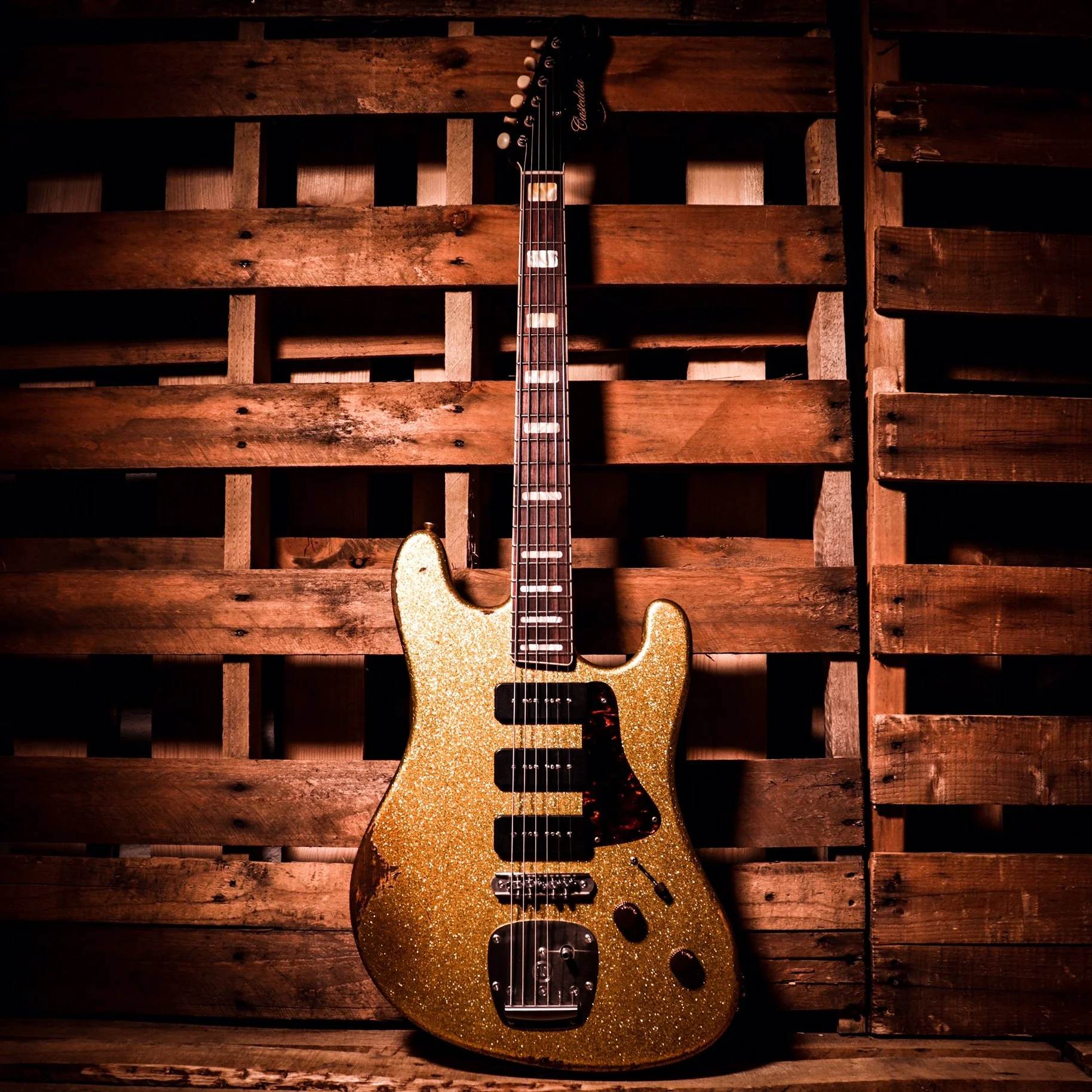 A gold glittery electric guitar with black pickups and a dark pickguard, leaning against a wooden pallet wall.