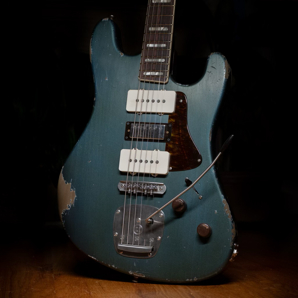 Vintage electric guitar with a worn blue finish, two white pickups, a dark pickguard, and a metal tremolo system, resting on a wooden surface.