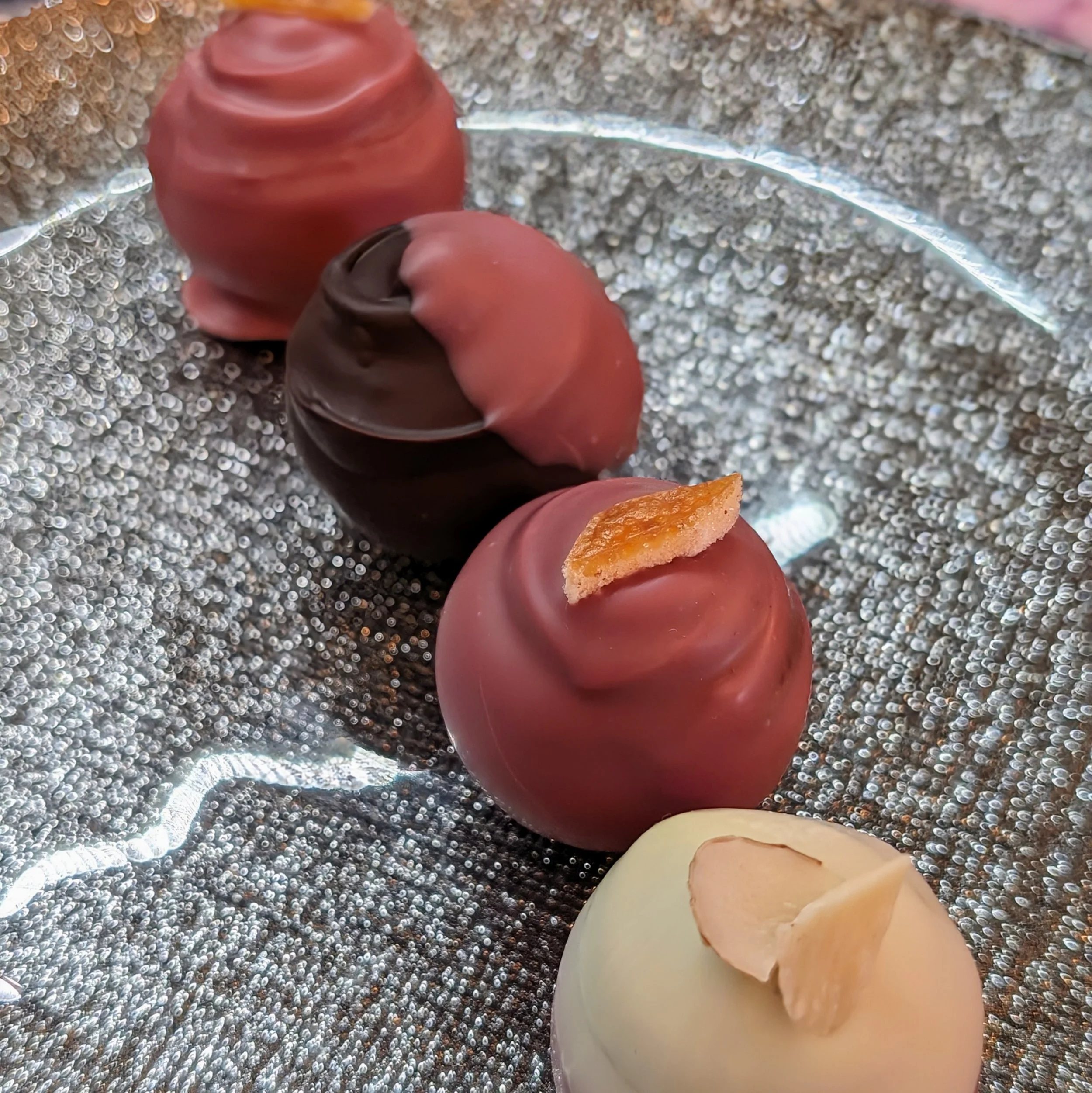 Four truffle chocolates with pink, dark, and white coatings, each topped with a small piece of candied fruit or nut, arranged on a reflective, textured silver surface.