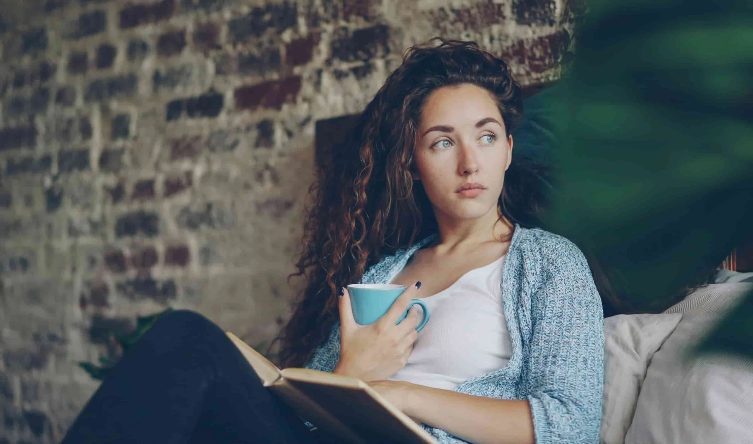 girl sitting in bed with a book and cup of coffee with head turned to the right thinking