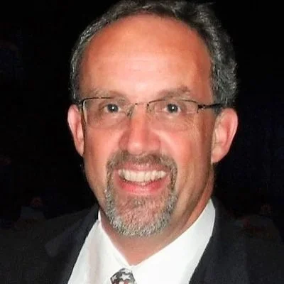 A man with glasses, short dark hair, and a beard wearing a tuxedo with a white shirt and patterned tie, smiling at the camera.