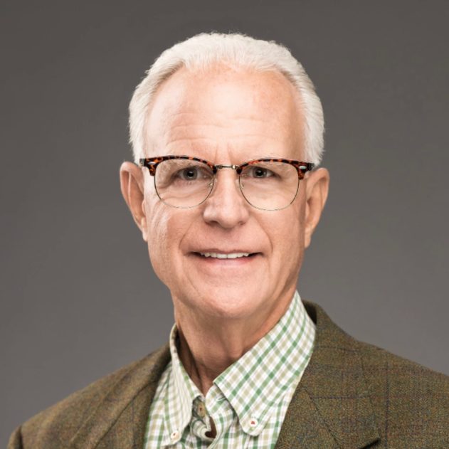A senior man with white hair, glasses with tortoiseshell frames, wearing a green and white checkered shirt and a brown jacket, smiling against a gray background.