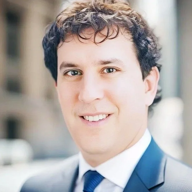 Headshot of a young man with dark and curly hair, wearing a suit and tie, smiling outdoors.