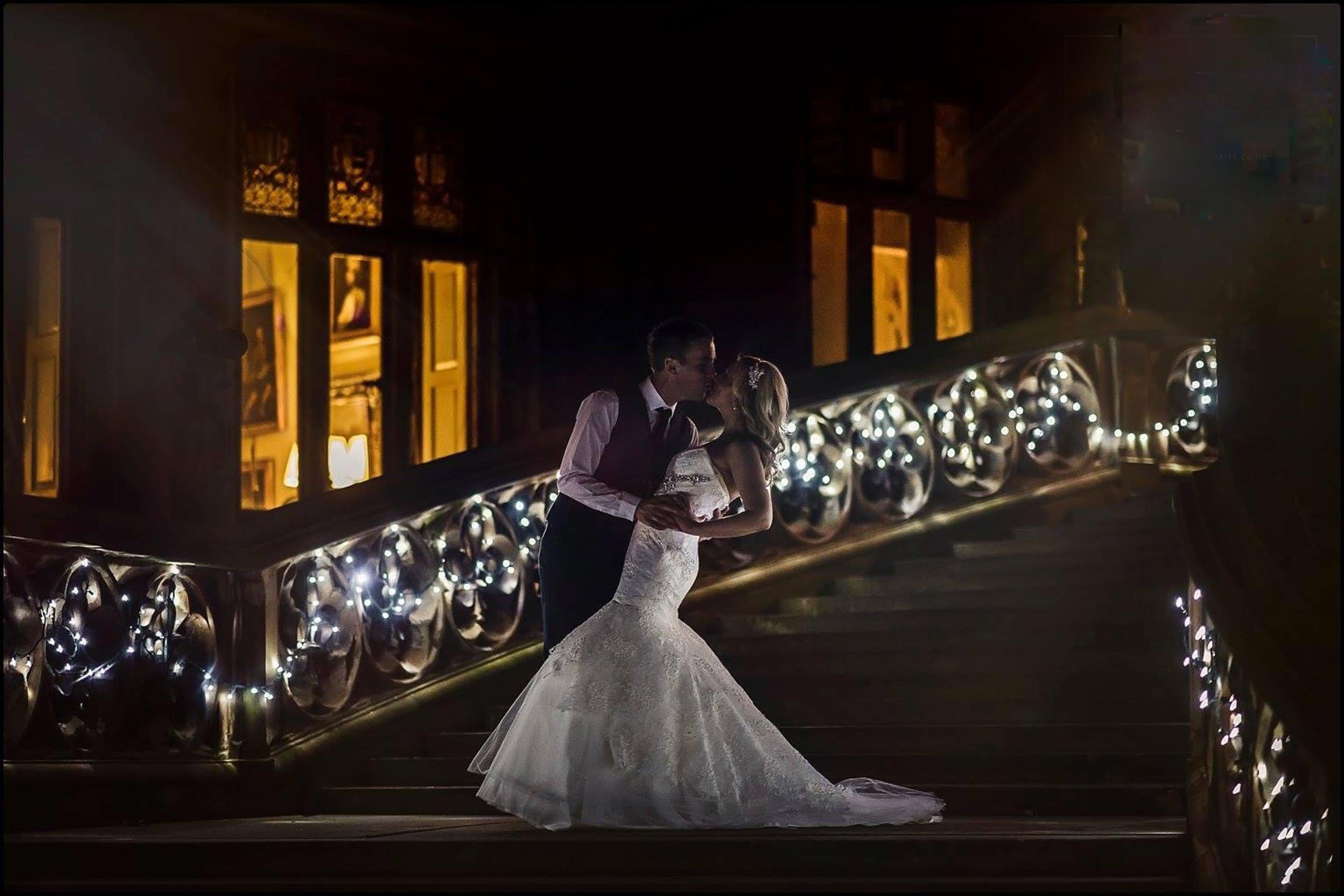 A bride and groom sharing a kiss on a staircase decorated with string lights at night in an elegant setting.