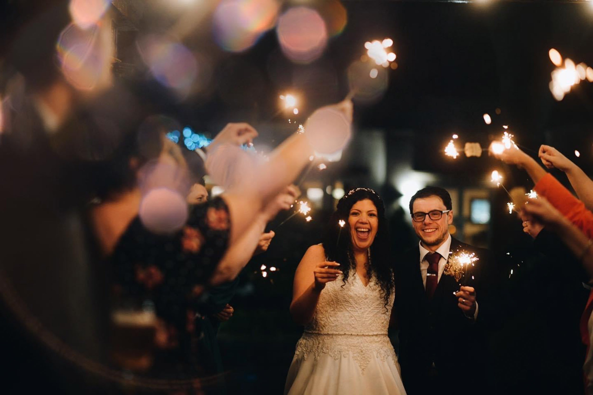 A bride and groom holding sparklers during their wedding celebration at night, surrounded by friends and family, with joyful expressions.