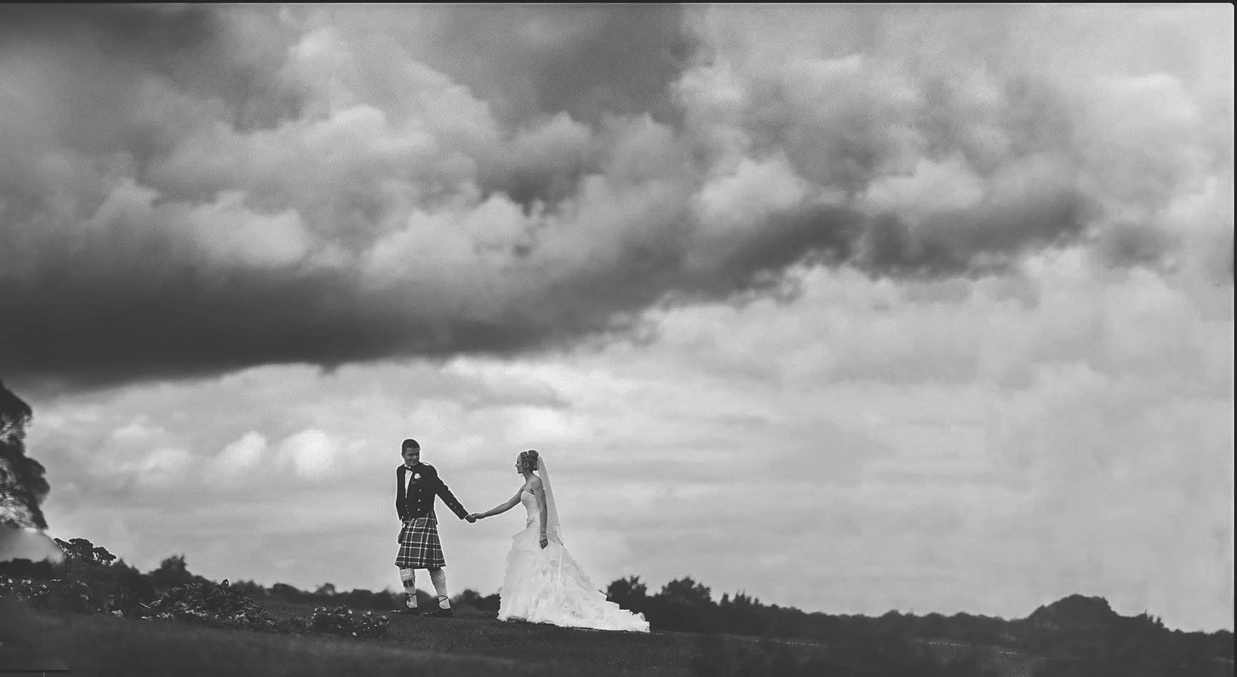 A black-and-white photo of a bride and groom holding hands outdoors under a cloudy sky, with trees in the background.