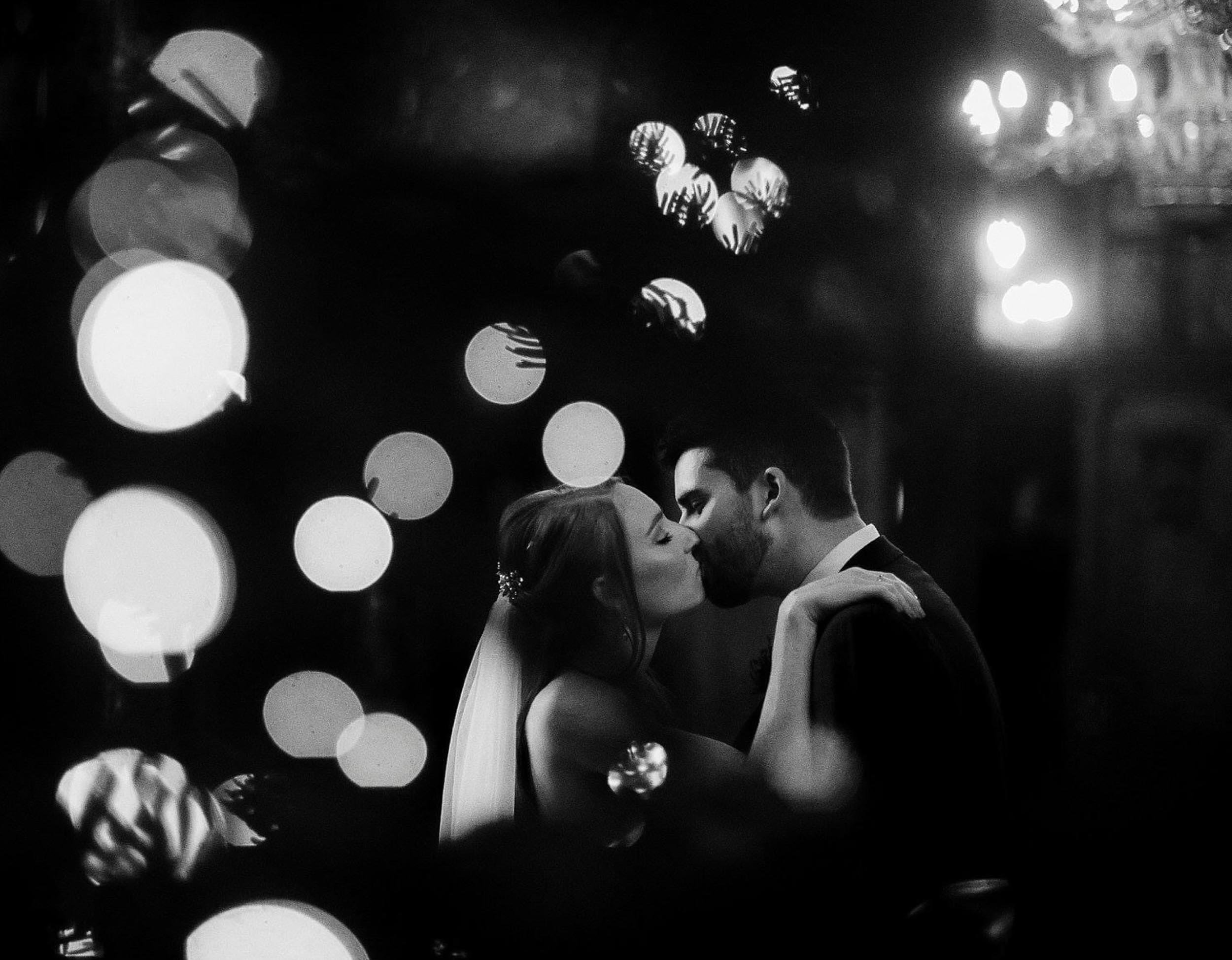 A black and white photograph of a bride and groom sharing a kiss, surrounded by out-of-focus lights creating a romantic atmosphere.