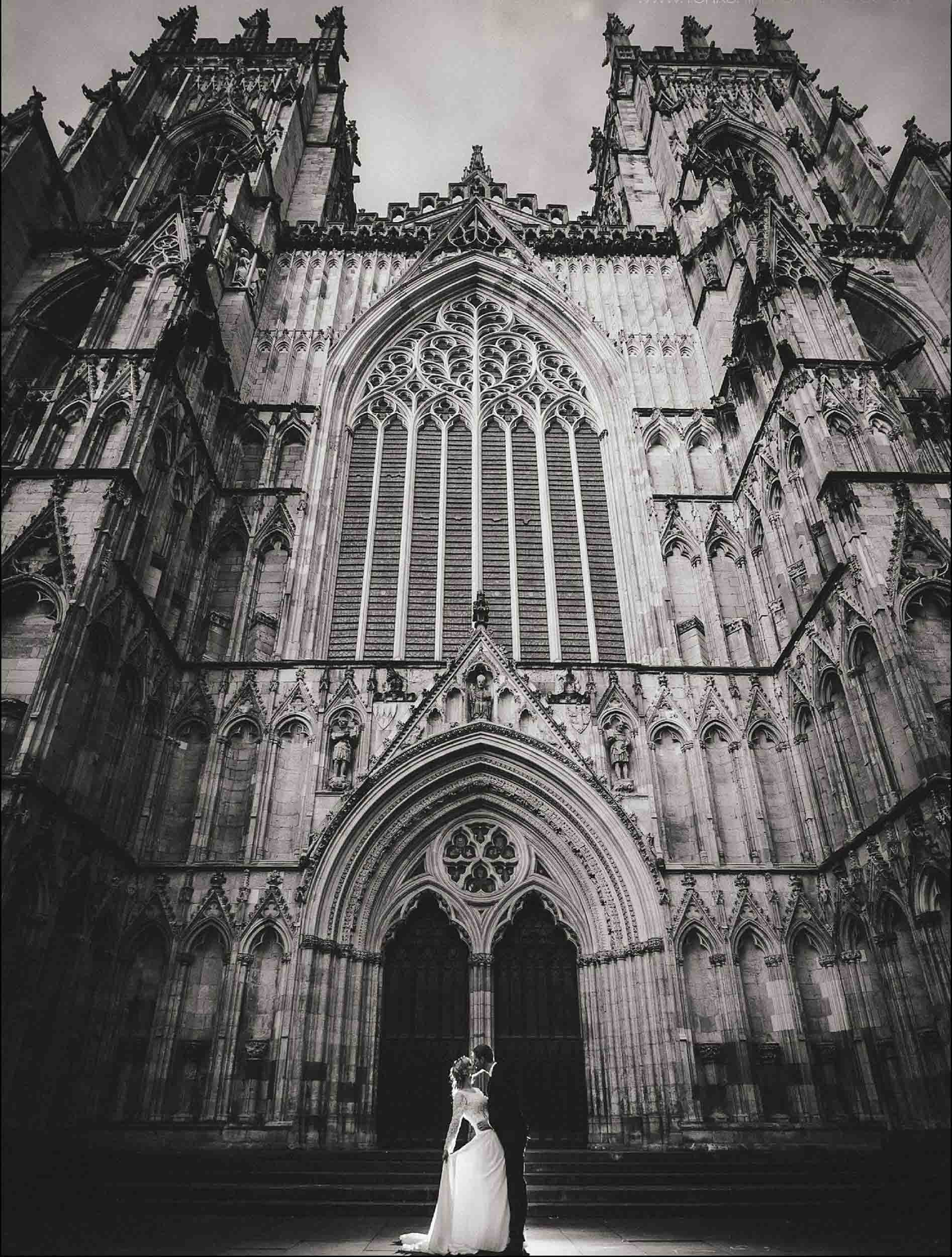A couple in wedding attire standing in front of a large, Gothic-style cathedral with detailed stone architecture and tall stained glass windows.
