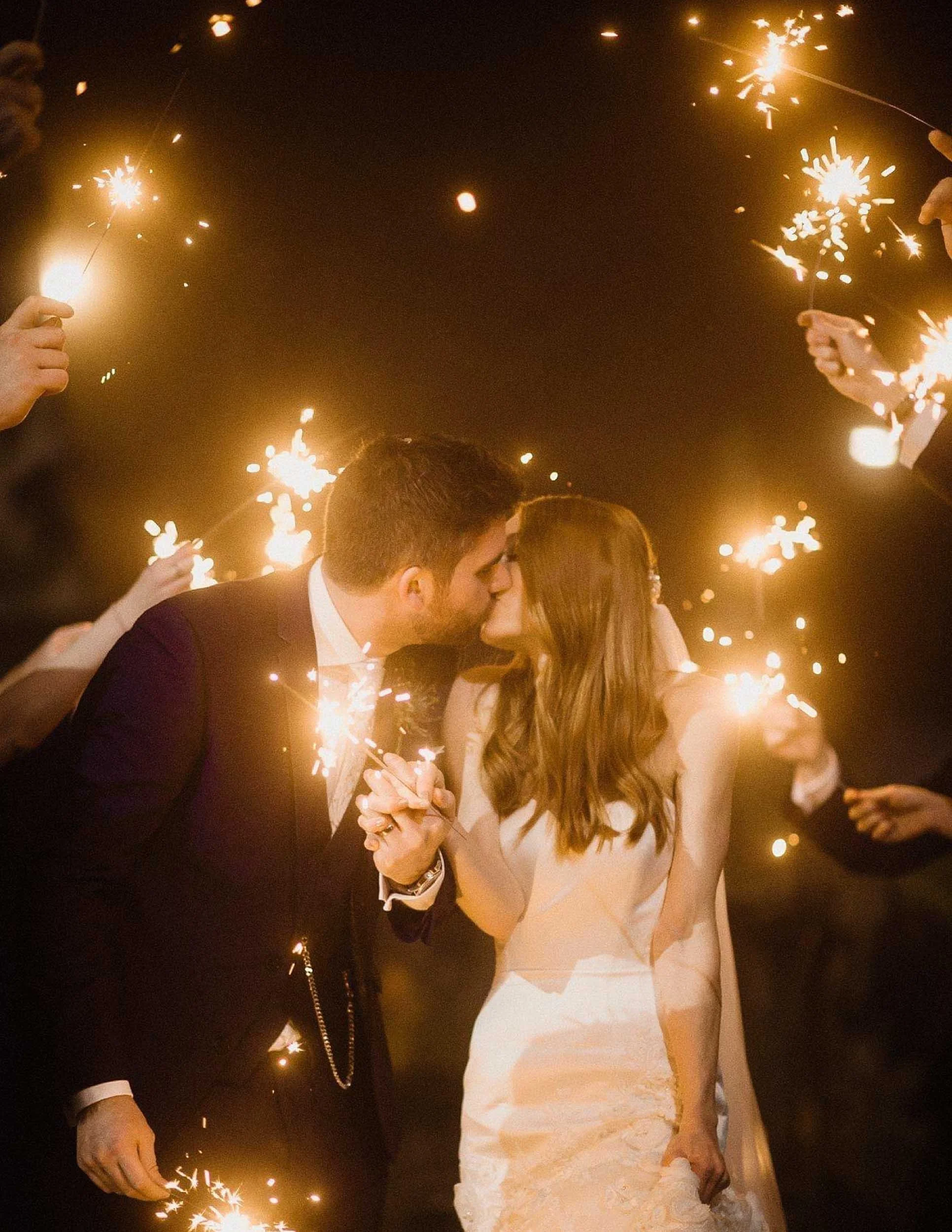 A couple in wedding attire sharing a kiss during a celebration with sparklers surrounding them at night.