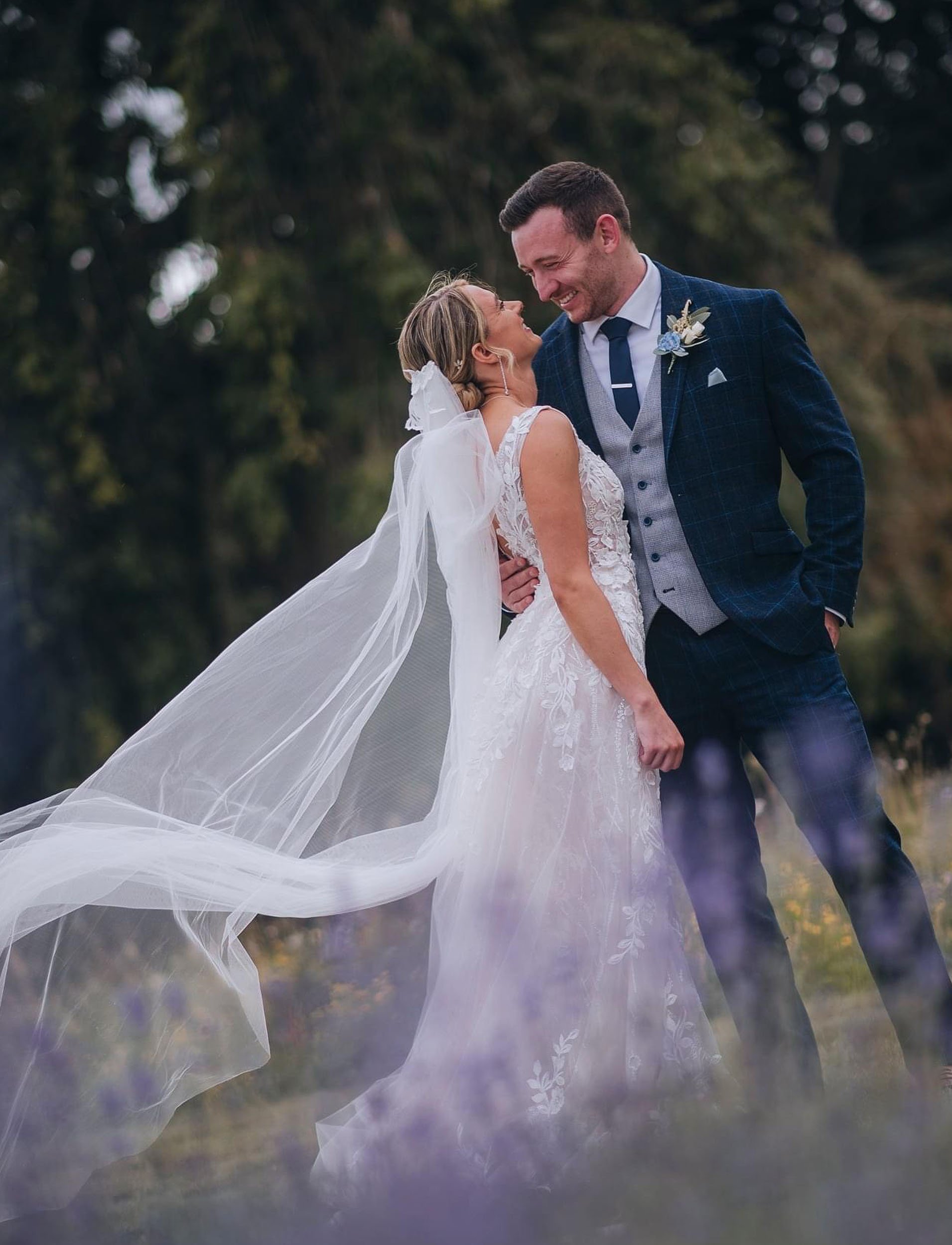 A bride and groom share a joyful moment outdoors, the bride wearing a lace wedding dress with a long veil, and the groom in a blue plaid suit, smiling and looking at each other.