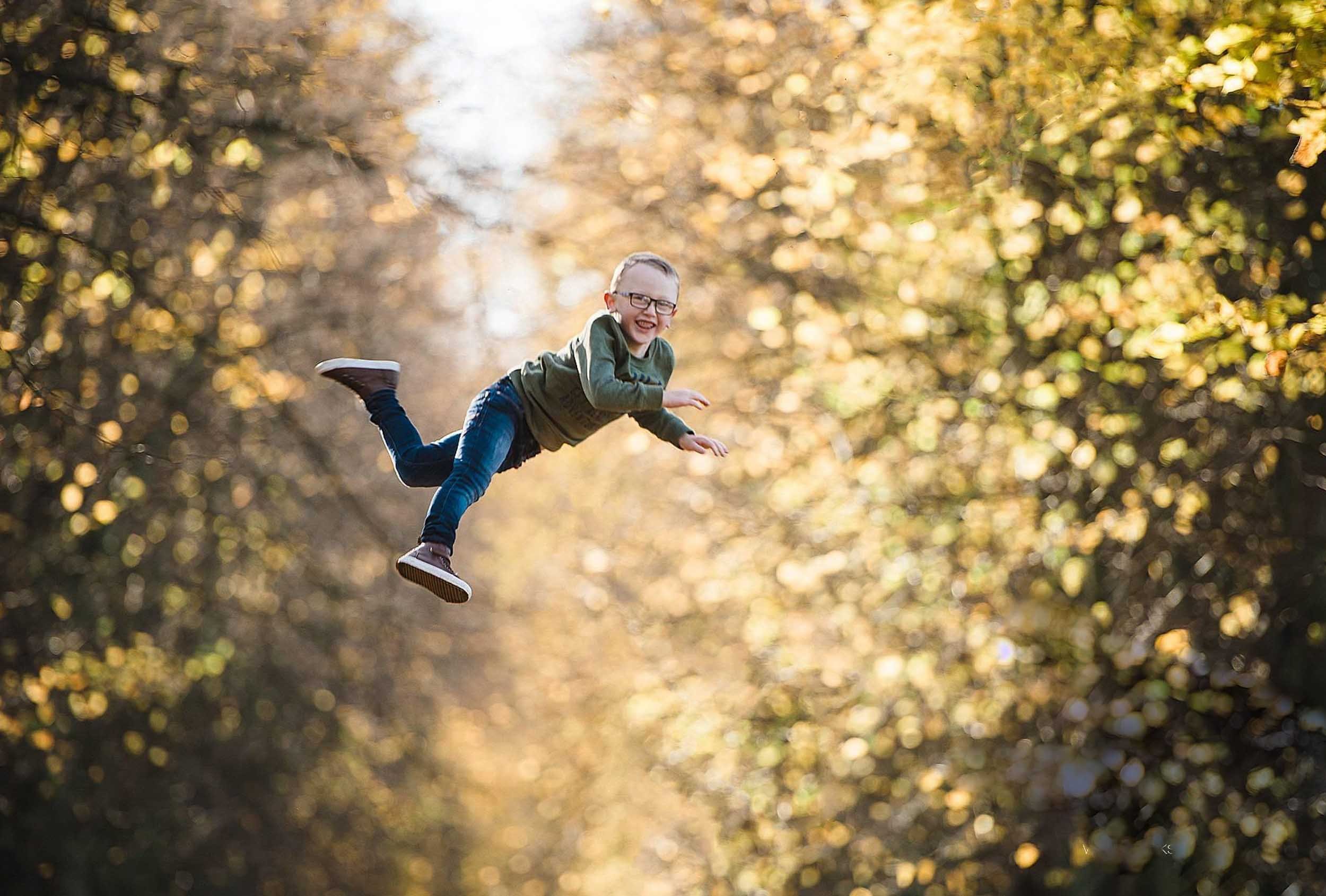A young boy with glasses, wearing a green shirt and jeans, smiling and falling through the air in a forest with autumn leaves.