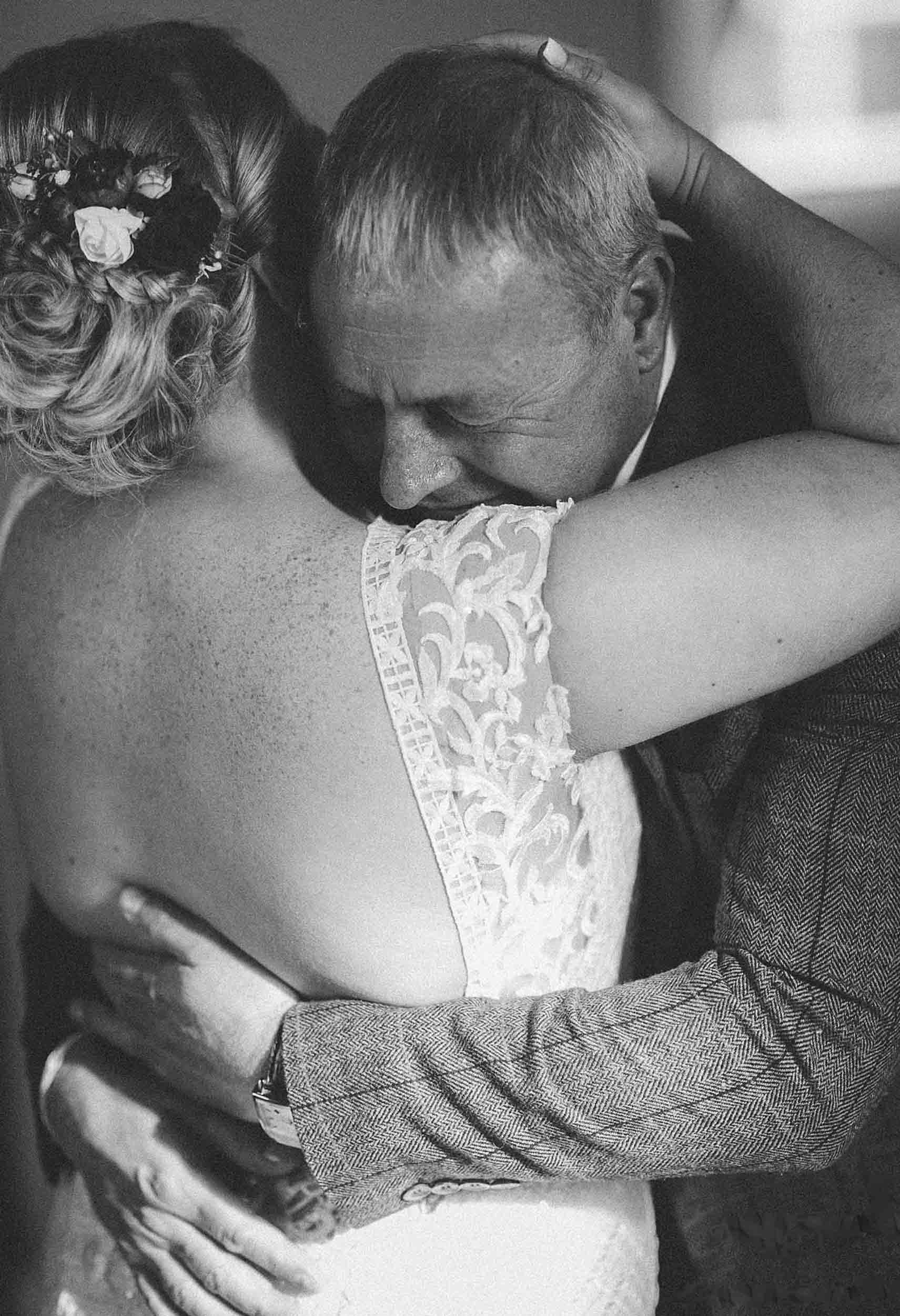 A black and white photo of a man and a woman embracing, with the man showing affection and the woman wearing a lace dress and floral hair accessory.