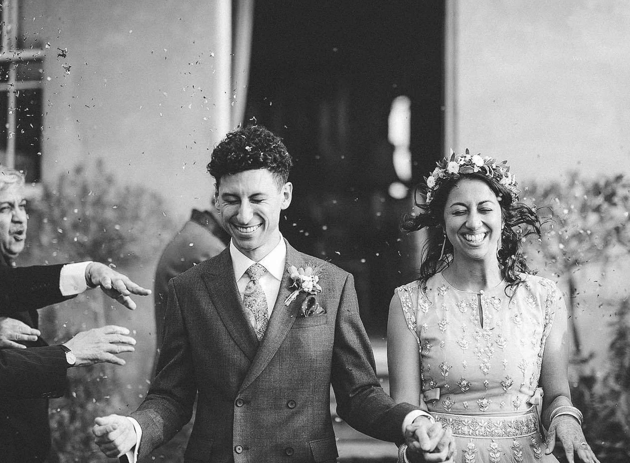 A black-and-white photo of a happy couple, dressed in wedding attire, holding hands and smiling as they walk through a confetti shower, surrounded by friends and family.