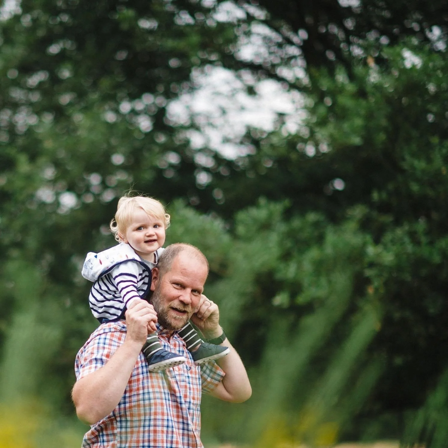 A man with a beard carrying a blonde young girl on his shoulders outdoors in a garden or park with green trees in the background. Family portrait in Clumber Park