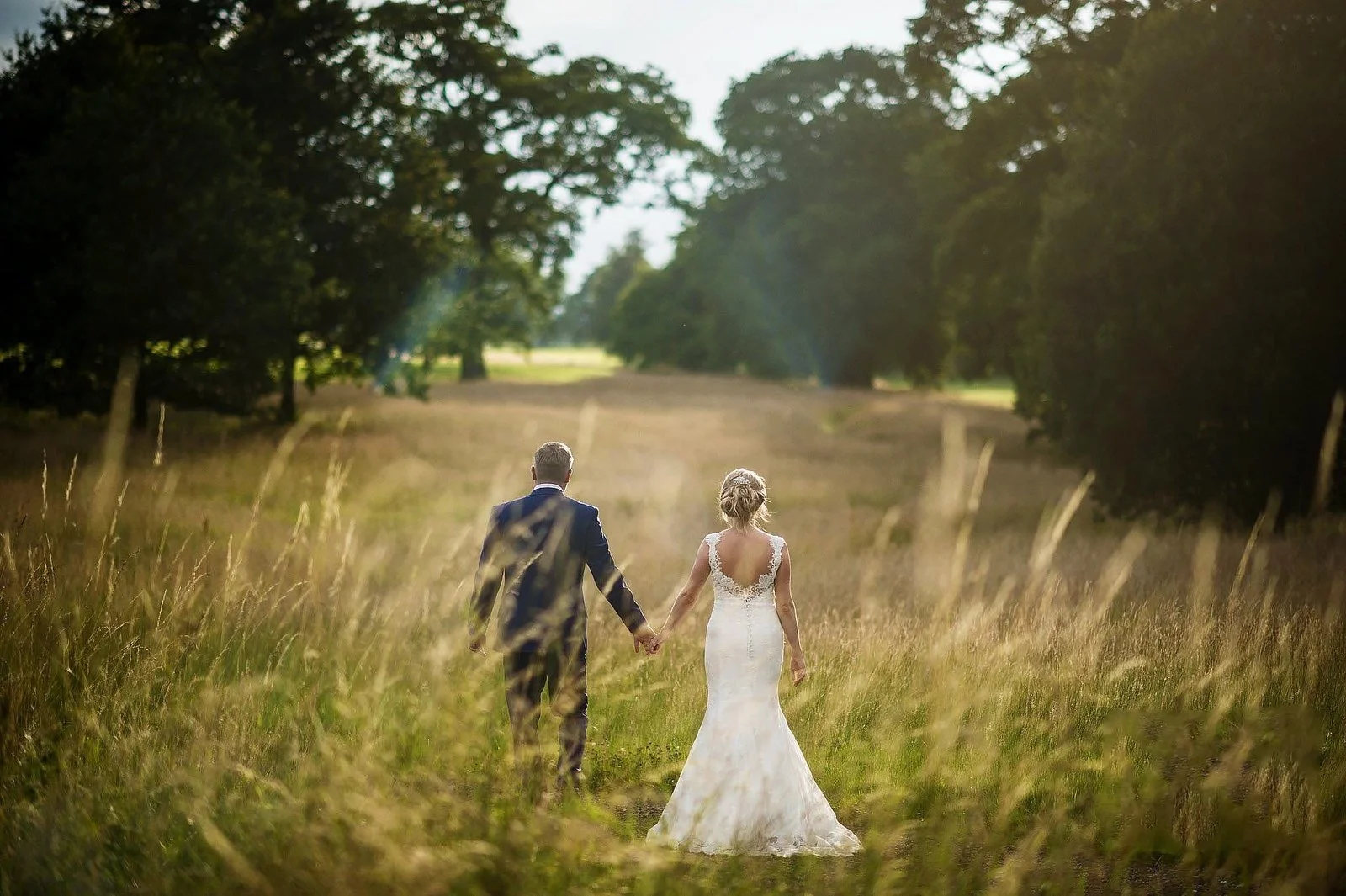 A bride and groom walking hand in hand through a grassy field surrounded by trees, with the bride wearing a white wedding gown and the groom in a dark suit.