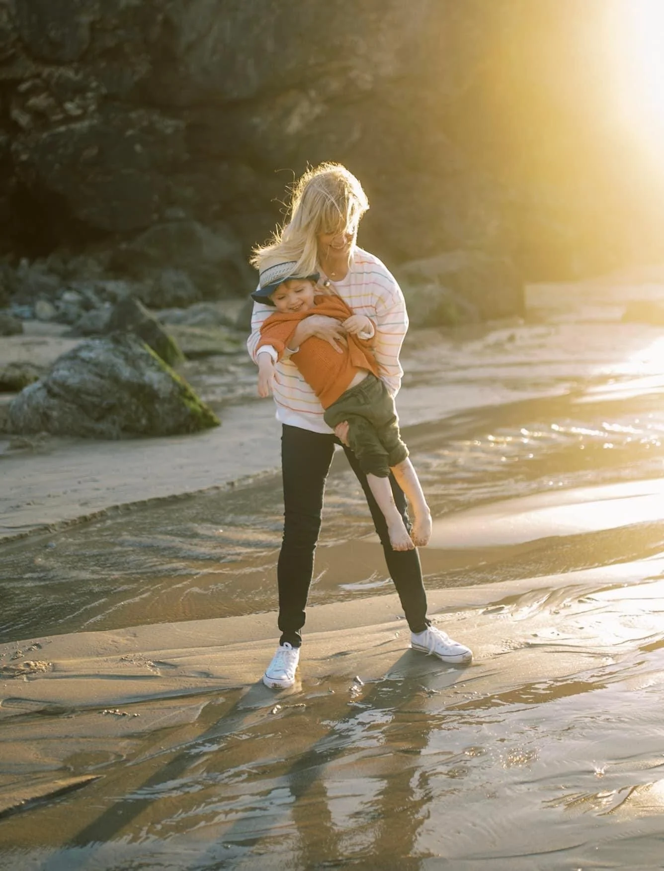 A woman playing with a young boy on a beach during sunset, lifting him in the air as they stand in shallow water near rocks.