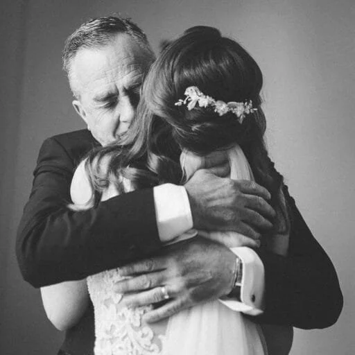A black-and-white photo captures the essence of Yorkshire wedding photography, with a bride and  father hugging tightly. The brides back reveals her lace dress and floral hairpiece, while the mans face is visible, eyes closed,  a tender expression.